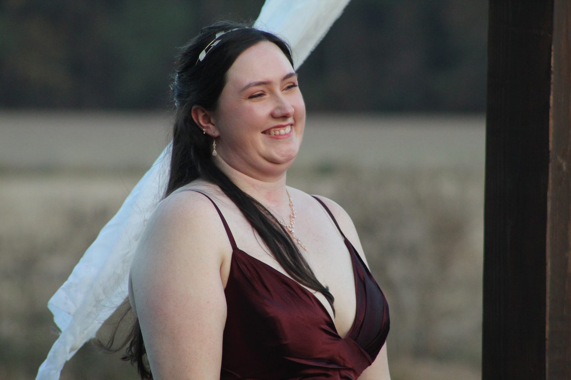 A smiling bridesmaid in a deep burgundy satin gown with a delicate necklace and headband laughs candidly against a soft blurred farmland backdrop at a historic estate wedding.