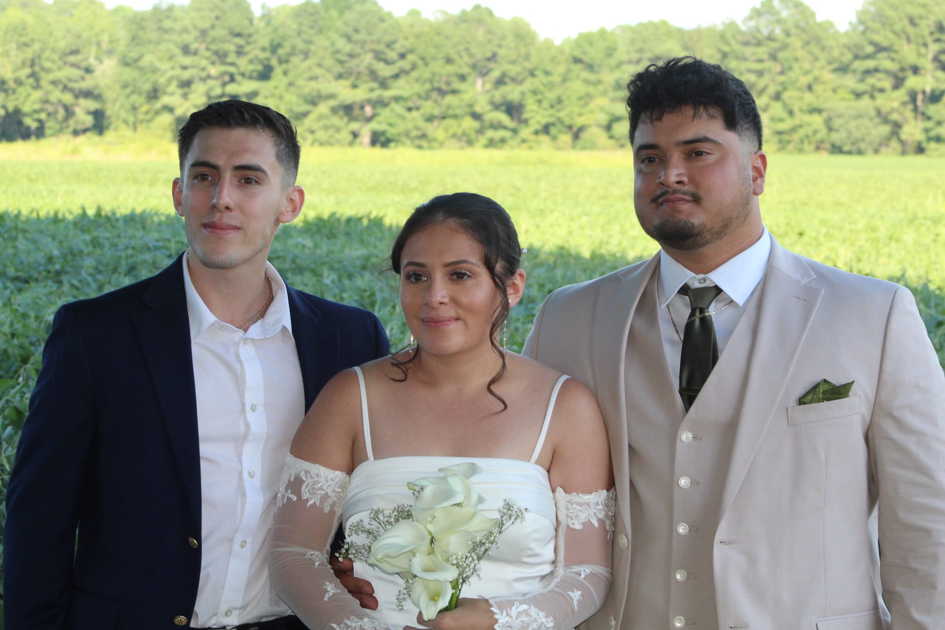 Bride in a white lace off-shoulder gown holding white calla lilies poses with two formally dressed guests in front of lush green farmland fields at Scarborough House.