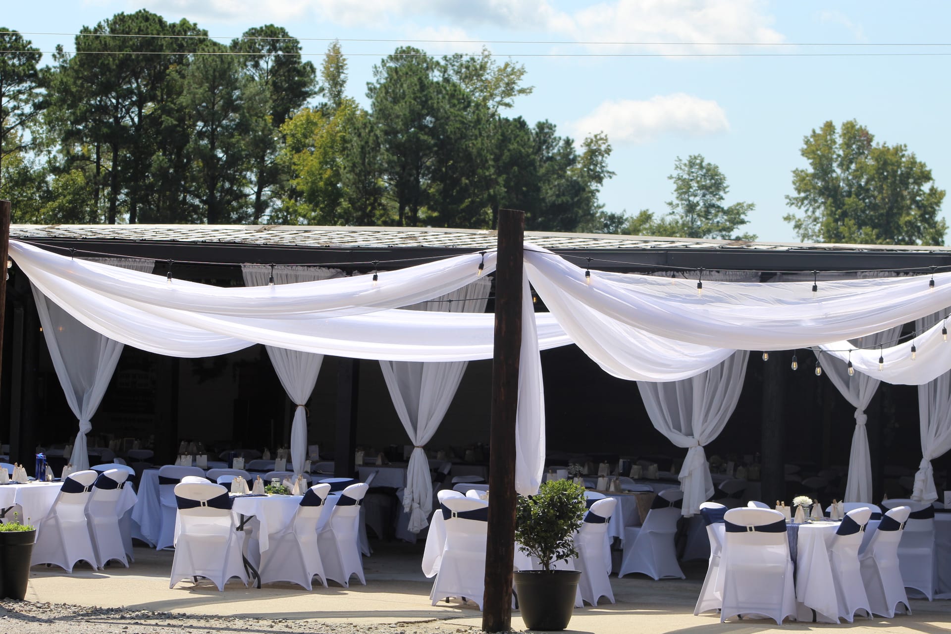 Outdoor wedding reception pavilion at Scarborough House elegantly dressed with sweeping white fabric draping, string lights, and round tables set with white linens and navy chair sashes surrounded by lush trees.