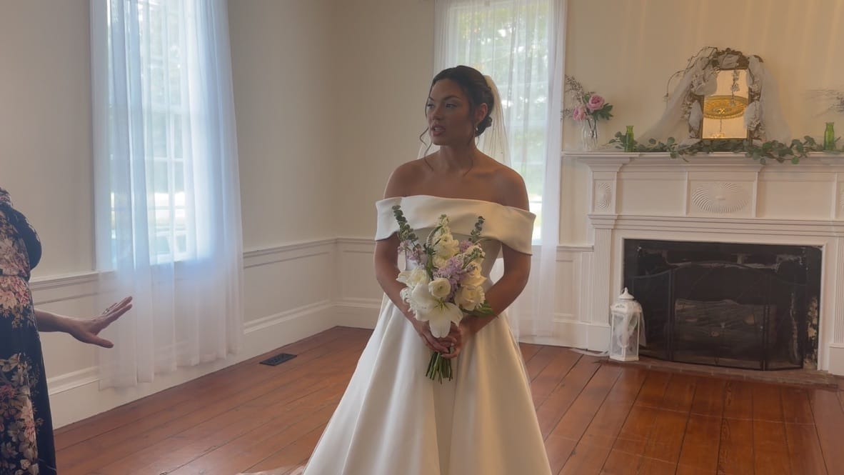 Bride in an off-shoulder white ball gown holding a pastel bouquet stands in the grand hall of Scarborough House with a decorated fireplace mantel visible behind her.