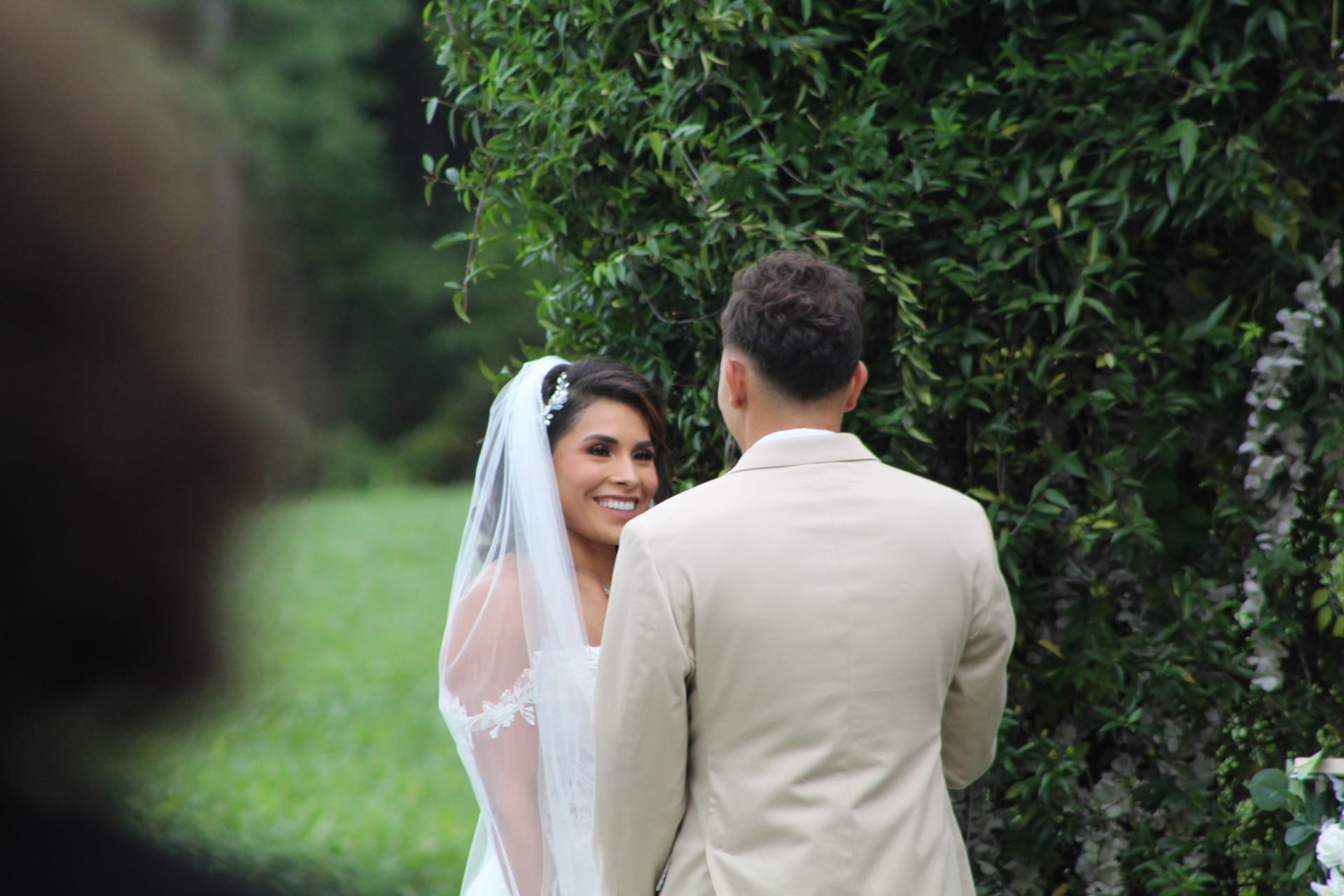 A beaming bride in a white gown and veil smiles lovingly at her groom during an outdoor garden ceremony at Scarborough House, framed by a lush green hedge backdrop.