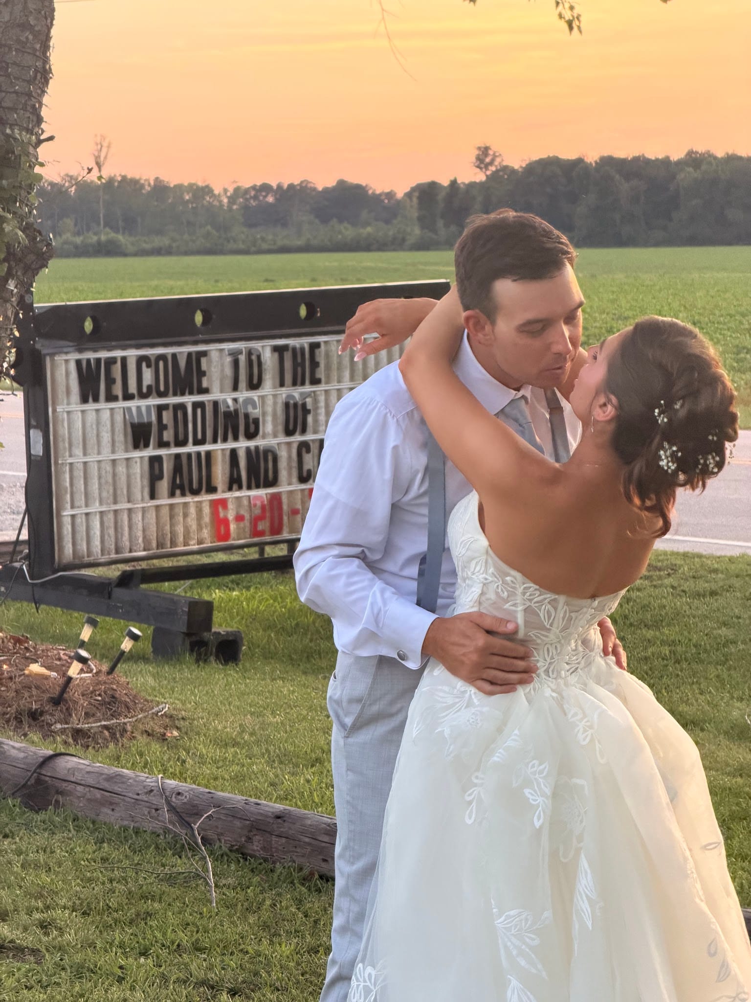 Bride and groom share a romantic kiss in front of a lit marquee welcome sign at sunset, with lush green farmland fields and a warm golden sky in the background.