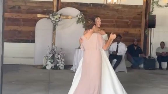 Bride in a white gown dances with a woman in a blush pink dress inside a rustic wood-paneled reception hall decorated with floral arrangements and an arched backdrop.