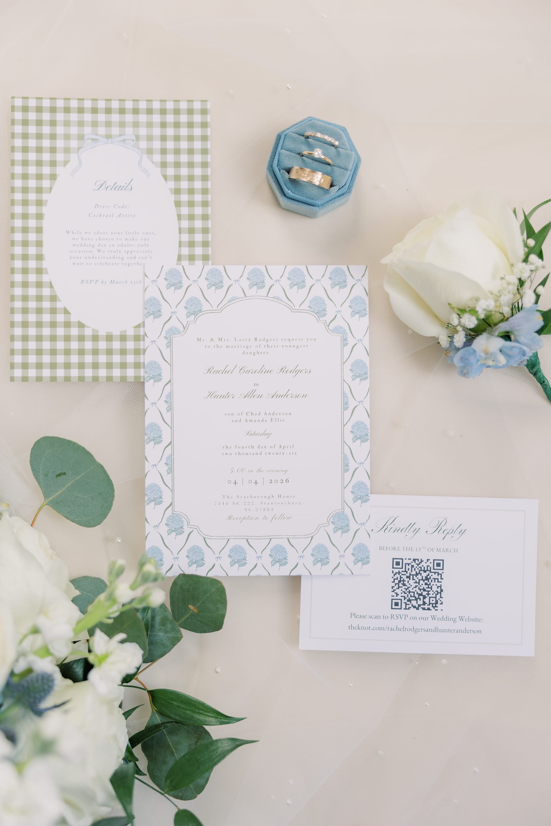 Elegant wedding invitation suite featuring blue floral stationery, gingham details card, QR code RSVP card, velvet ring box with gold bands, and white and blue floral accents arranged on a neutral surface.
