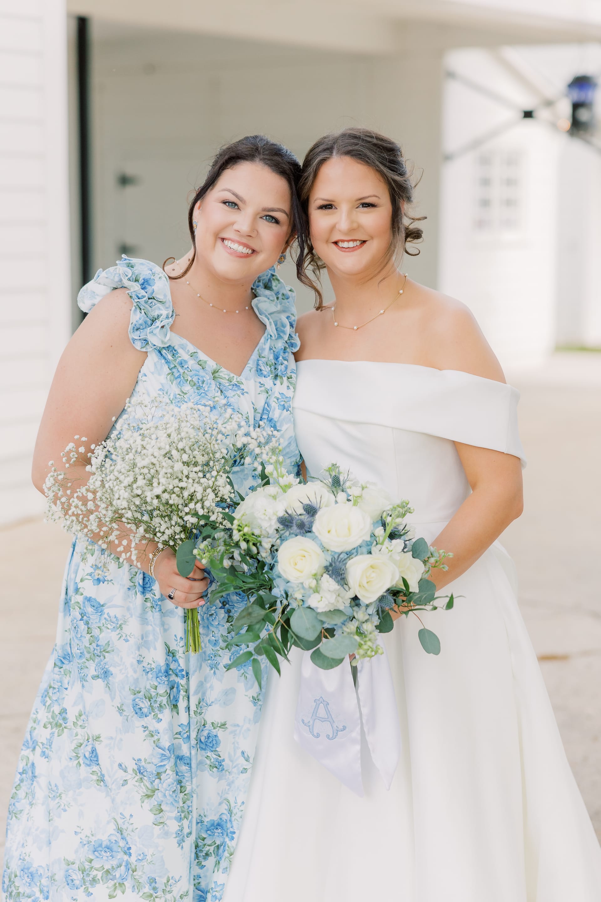 Bride in an off-shoulder white gown holding a white and blue floral bouquet poses with her bridesmaid in a blue floral dress holding a baby's breath bouquet outside Scarborough House.