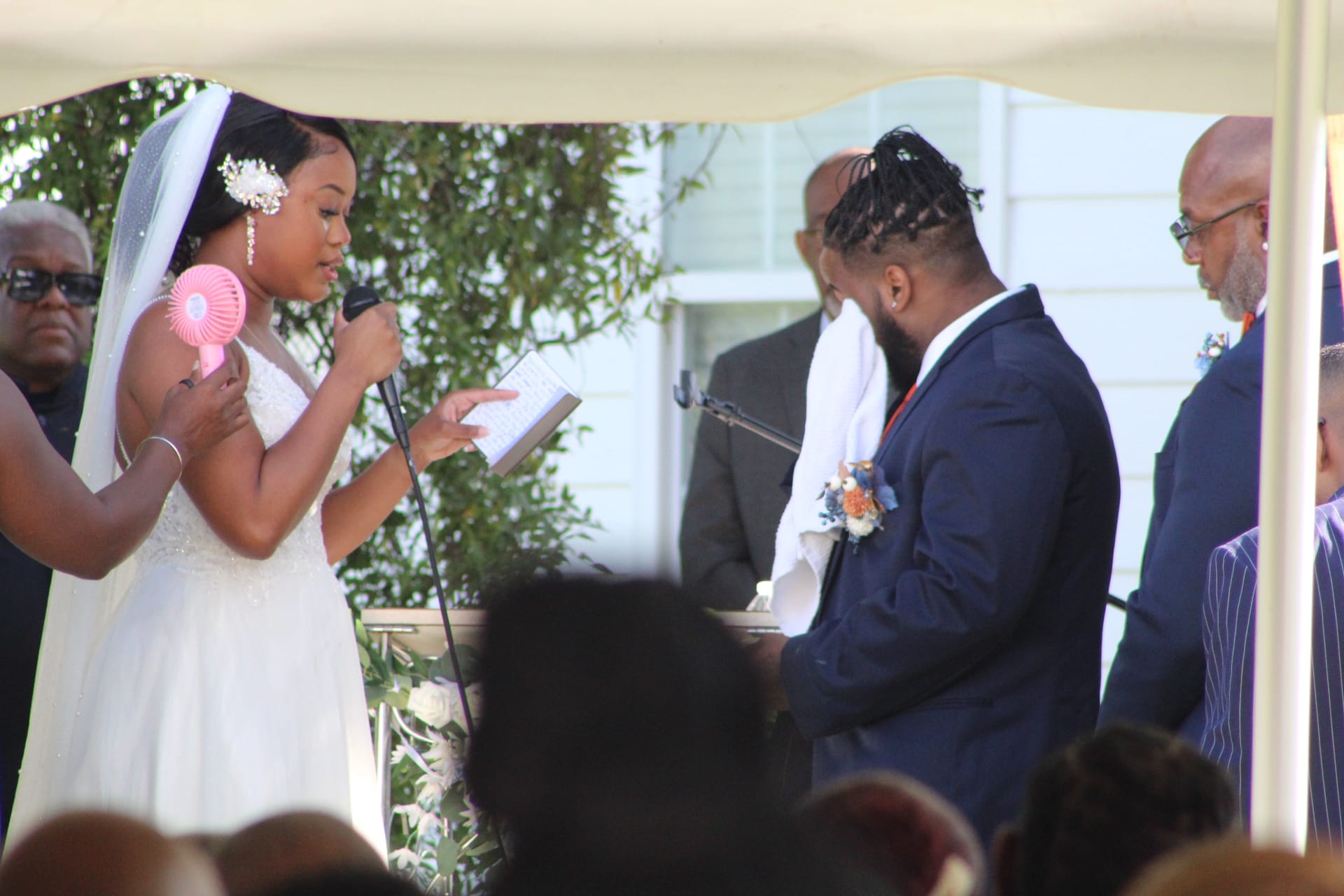 Bride reads her wedding vows from a book at an outdoor microphone while the groom bows his head emotionally during a sunny ceremony at a historic estate venue.