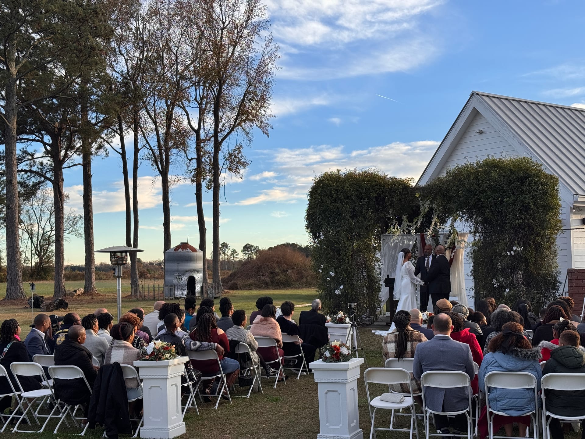 Outdoor wedding ceremony at Scarborough House with guests seated in white folding chairs facing a couple exchanging vows beneath a lush floral arch beside the historic white chapel, with tall pines and farmland fields in the background under a blue sky.