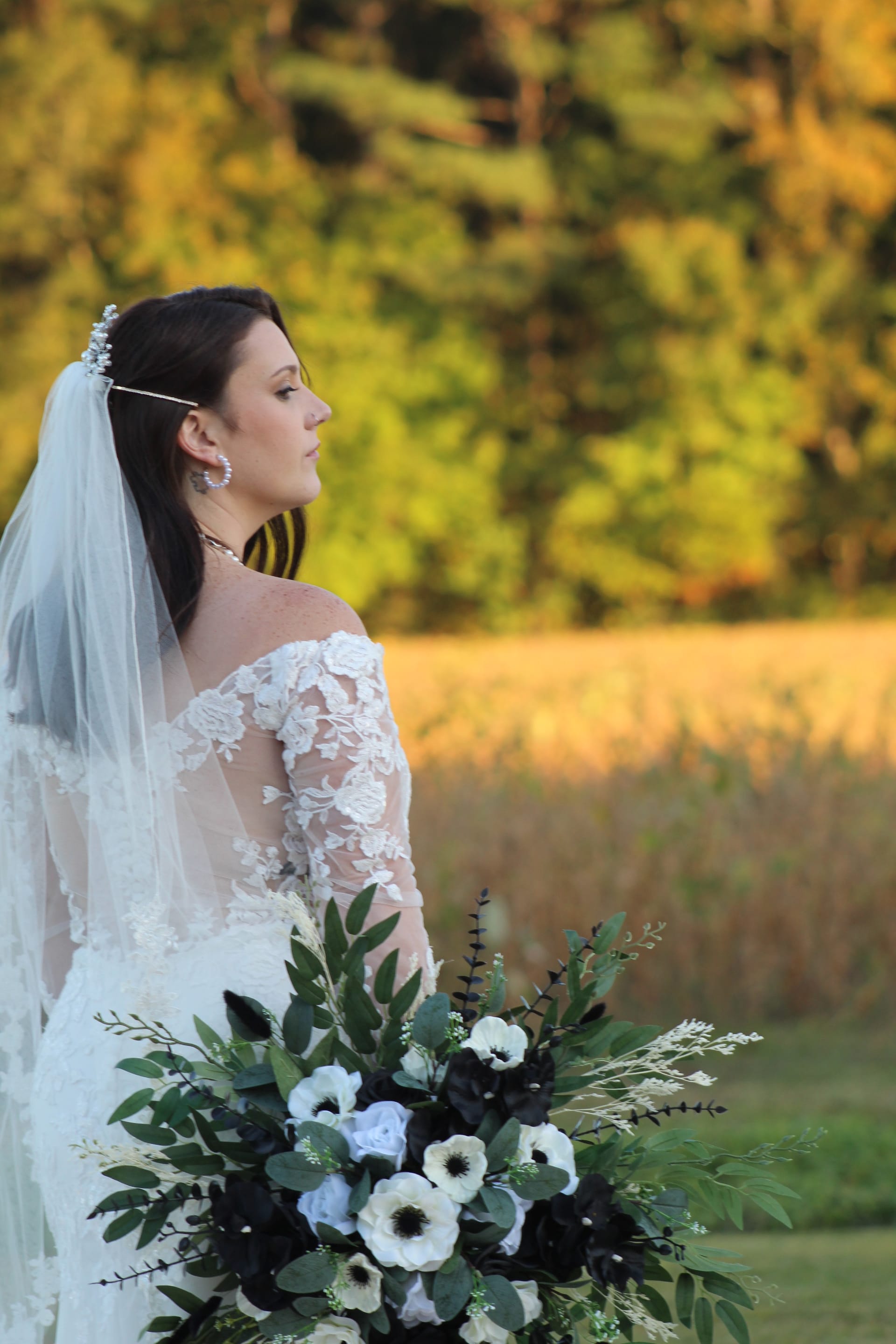 Bride in a lace off-shoulder gown holding a black and white floral bouquet gazes across golden farmland fields at sunset during a fall wedding at Scarborough House.