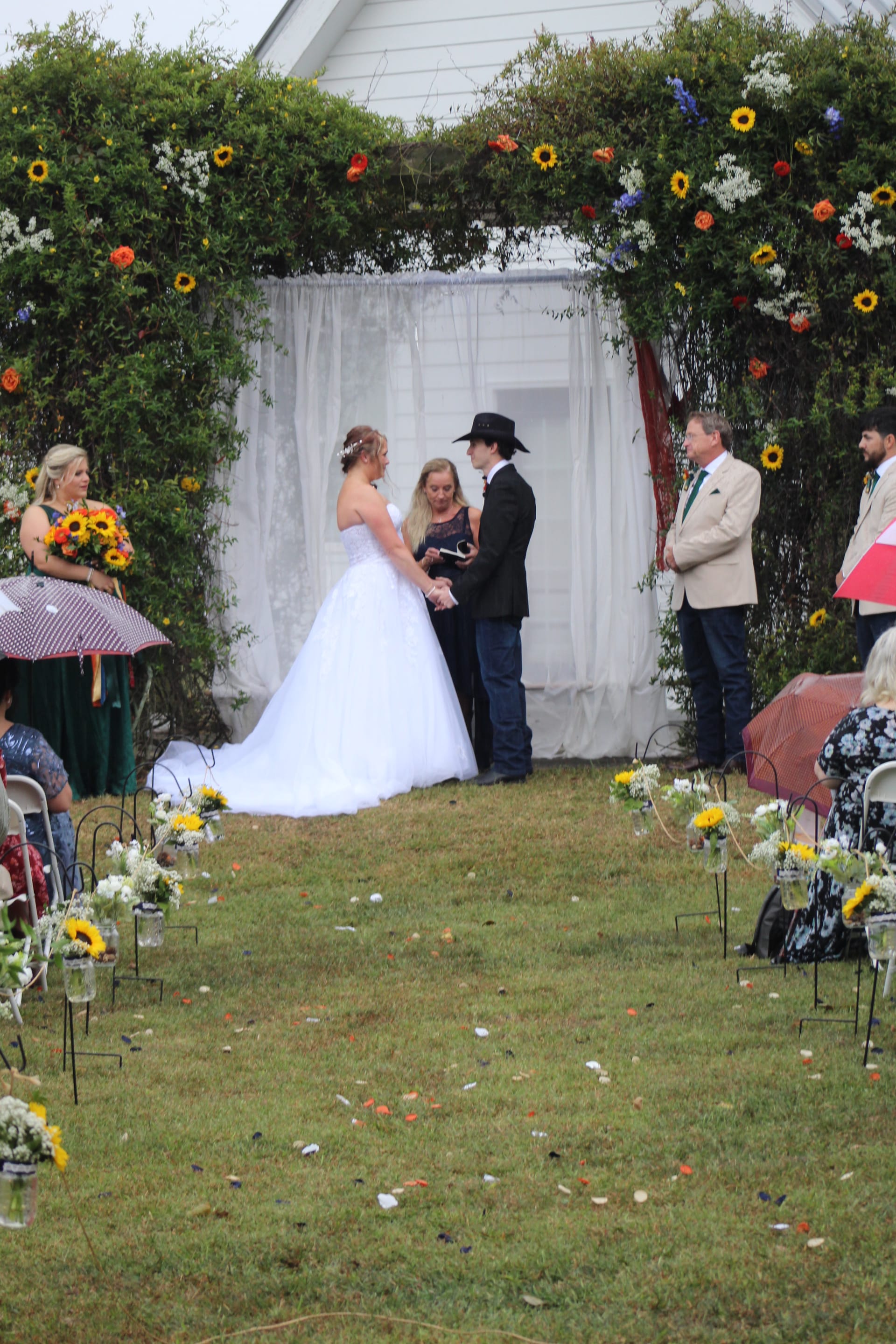 Bride in a white ball gown and groom in a black cowboy hat exchange vows beneath a lush greenery arch adorned with sunflowers and colorful blooms on the ceremony lawn at Scarborough House.