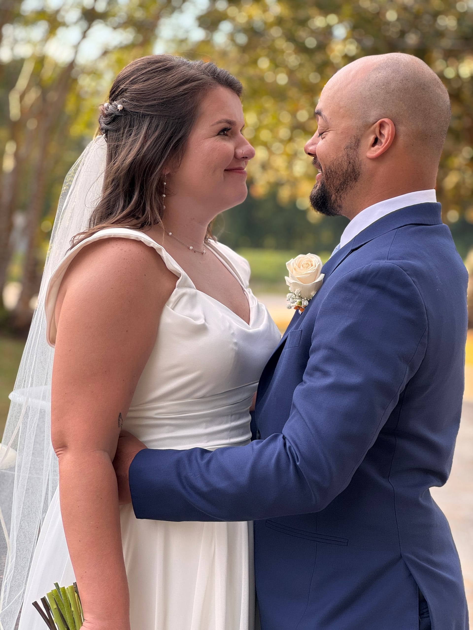 Bride in a white satin gown and veil and groom in a navy suit with a cream rose boutonniere share a tender smile while facing each other outdoors among soft-focused trees at Scarborough House.