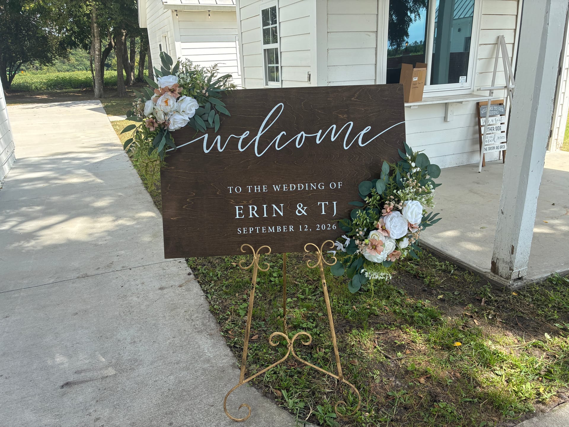 Rustic dark wood wedding welcome sign reading Welcome to the Wedding of Erin and TJ September 12 2026 adorned with white and blush floral arrangements on a gold ornate easel outside a white historic estate.