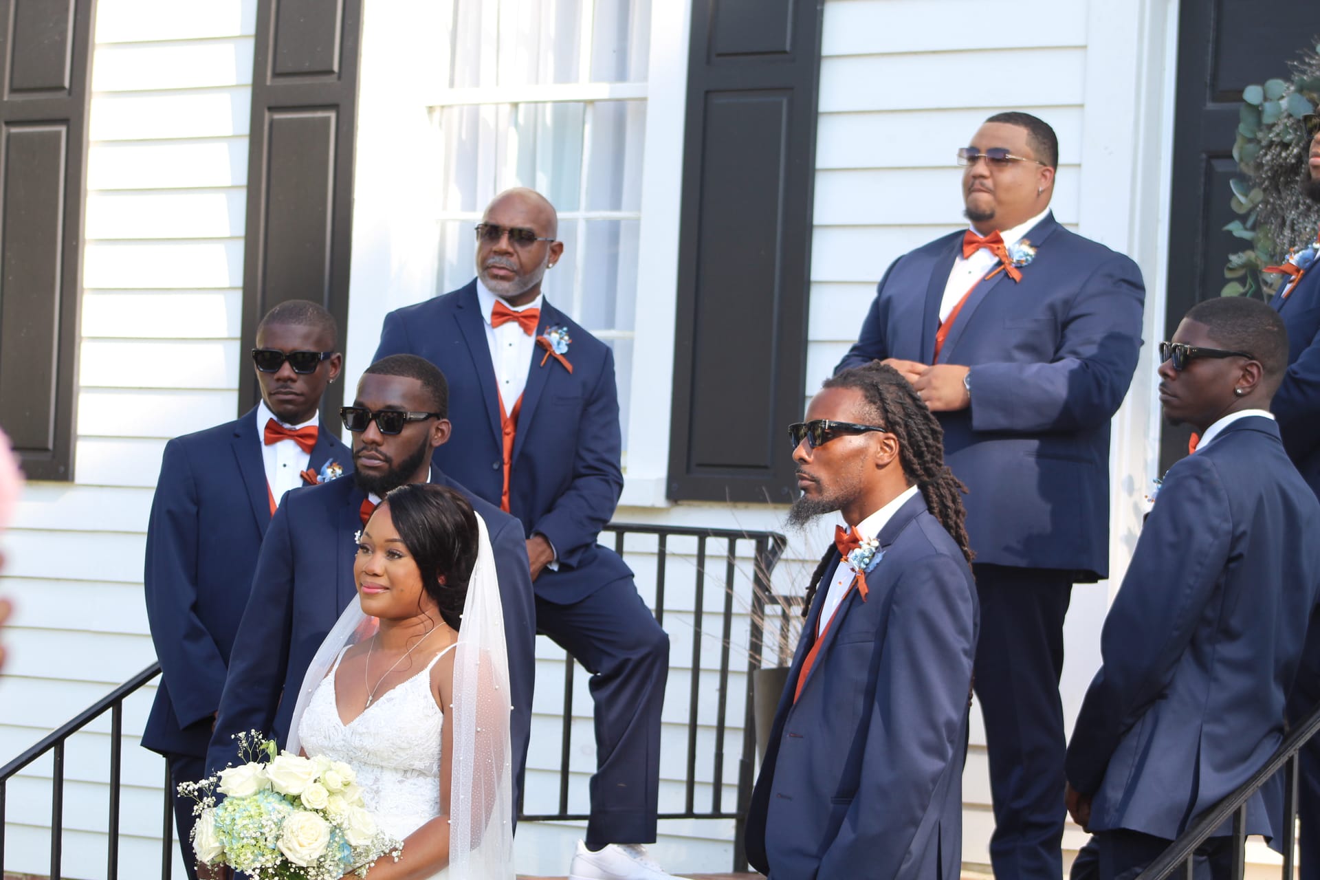 Bride in white gown holding a bouquet stands with groomsmen in navy suits and orange bow ties on the front steps of the historic Scarborough House estate.