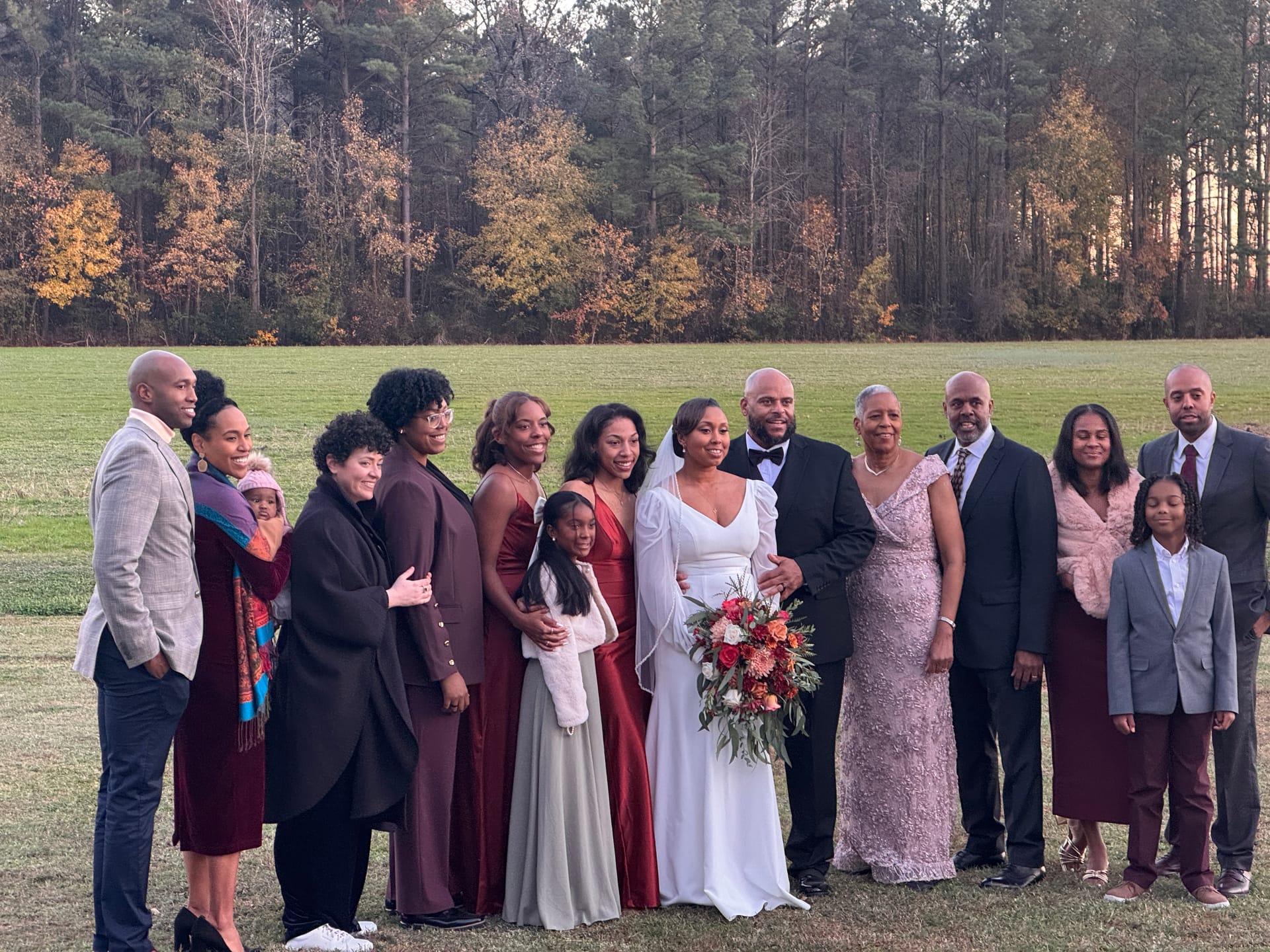 Bride in white gown and groom in black tuxedo pose with their wedding party and family on a sunlit ceremony lawn surrounded by autumn trees at Scarborough House estate.