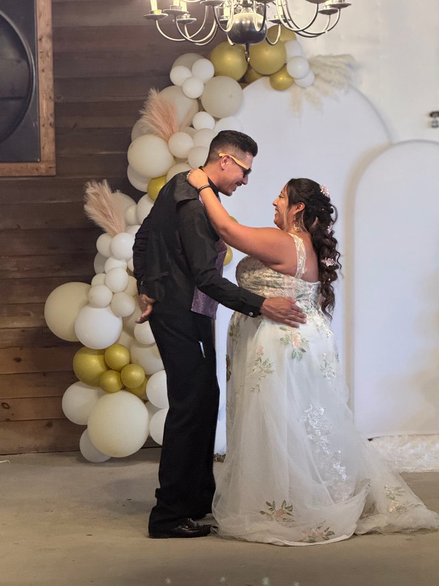 Bride in a floral white gown and groom in black share a joyful first dance in front of a gold and ivory balloon arch at Scarborough House.
