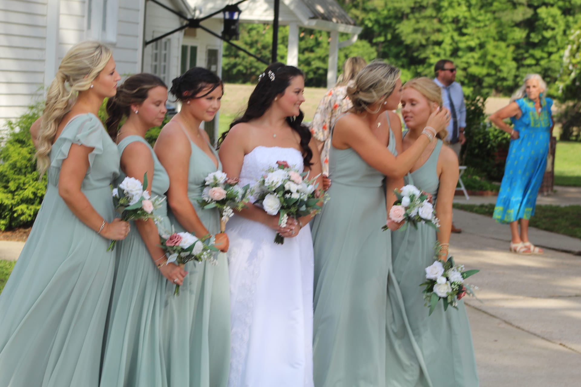 Bride in a white strapless gown shares an emotional moment with her bridesmaids in sage green dresses holding blush and white bouquets outside the historic Scarborough House manor.