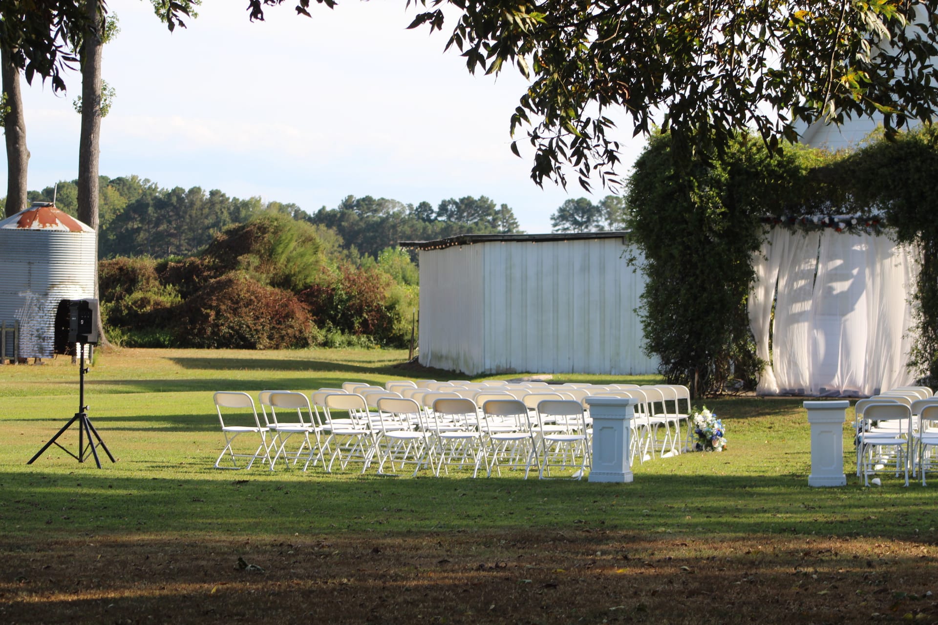 Outdoor wedding ceremony setup at Scarborough House featuring rows of white folding chairs, decorative white pillars with floral arrangements, a draped fabric arbor, and a rustic grain silo and barn in the background.