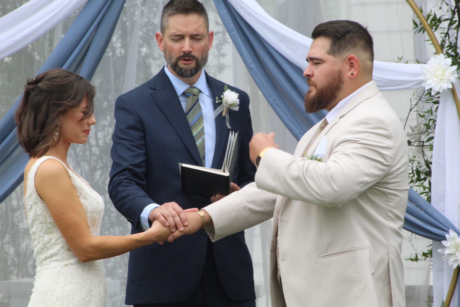 Groom placing a wedding ring on his brides hand during an outdoor ceremony at Scarborough House with a blue draped arch and white floral accents in the background.