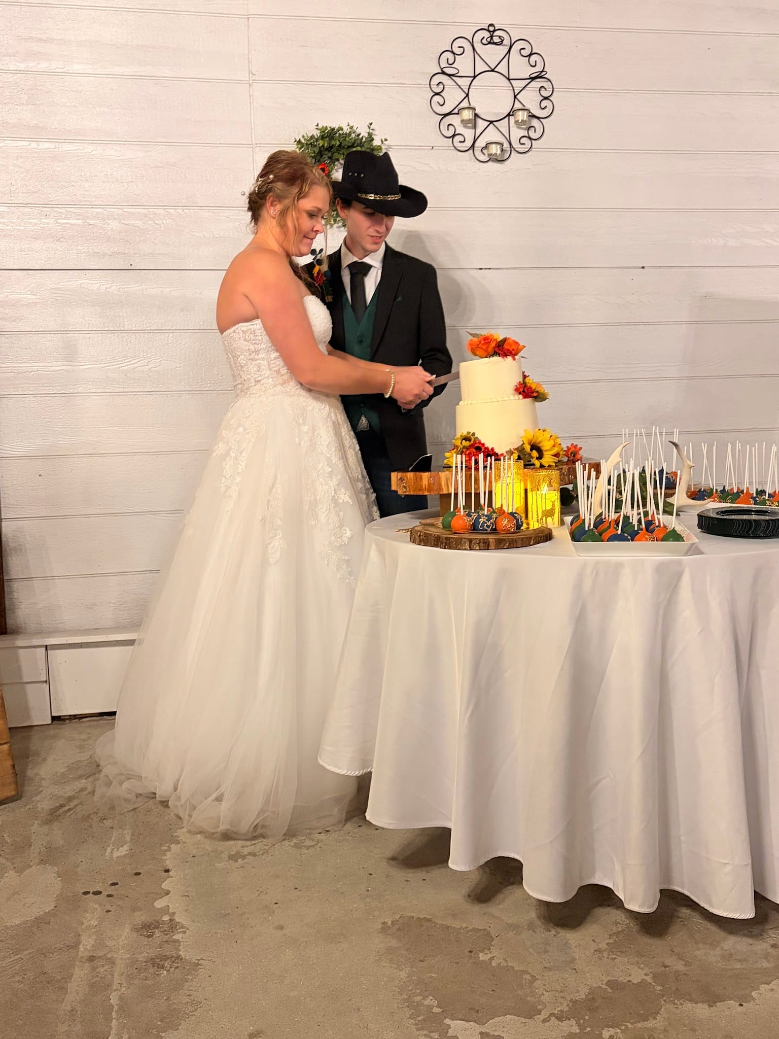 Bride in a white lace gown and groom in a black suit and cowboy hat cut their two-tier autumn-floral wedding cake at a rustic barn venue.