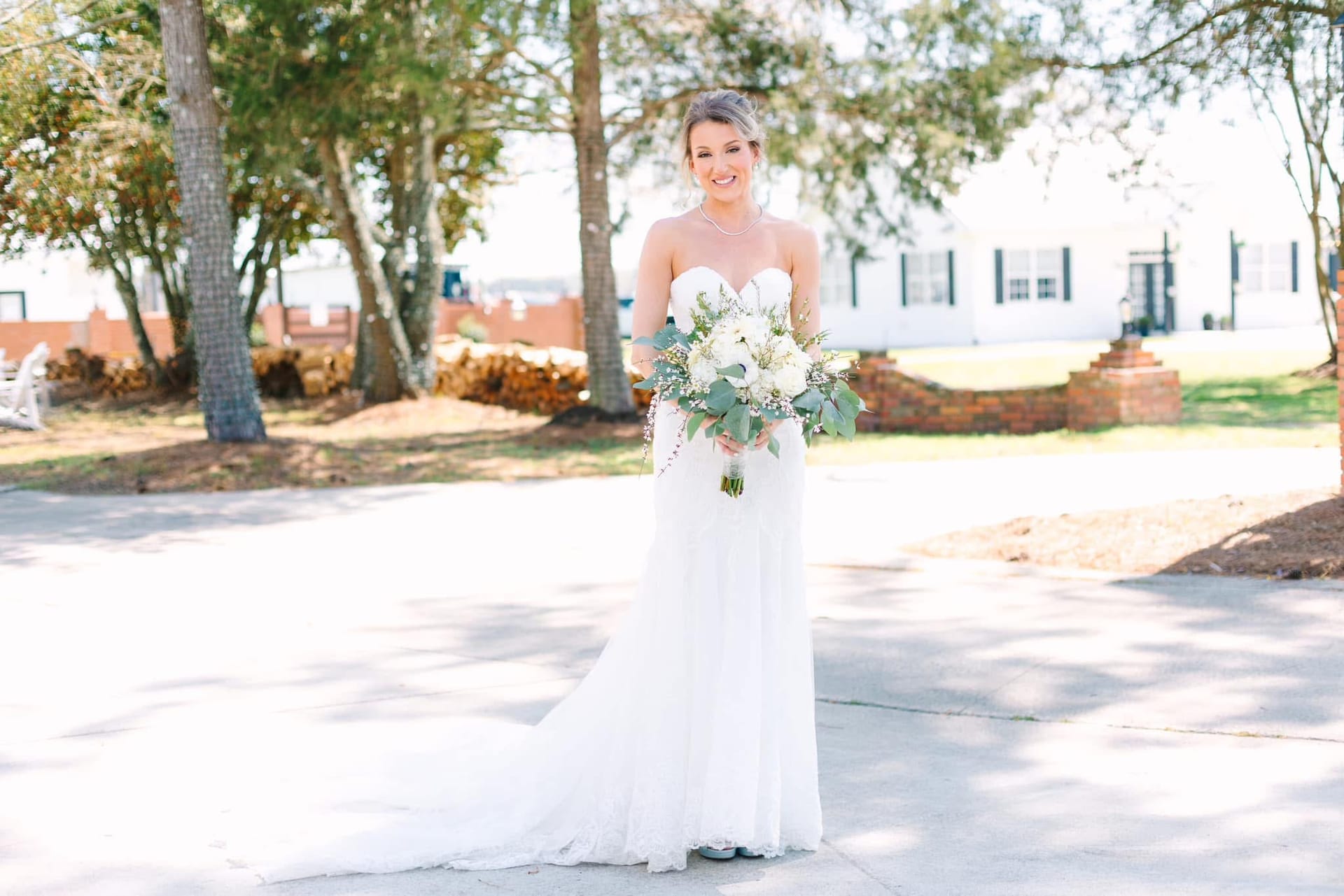 Radiant bride in a strapless white lace gown holding a white and greenery bouquet smiling in front of the historic 1821 Scarborough House manor surrounded by oak trees.