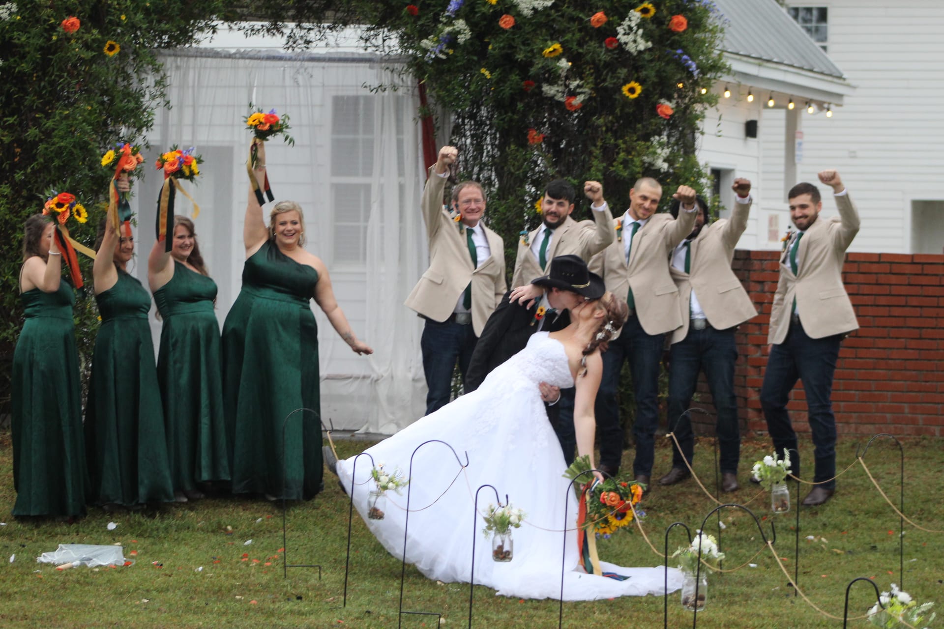 Bride and groom share a dramatic dip kiss on the ceremony lawn at Scarborough House while bridesmaids in emerald gowns raise bouquets and groomsmen in tan blazers cheer behind them.