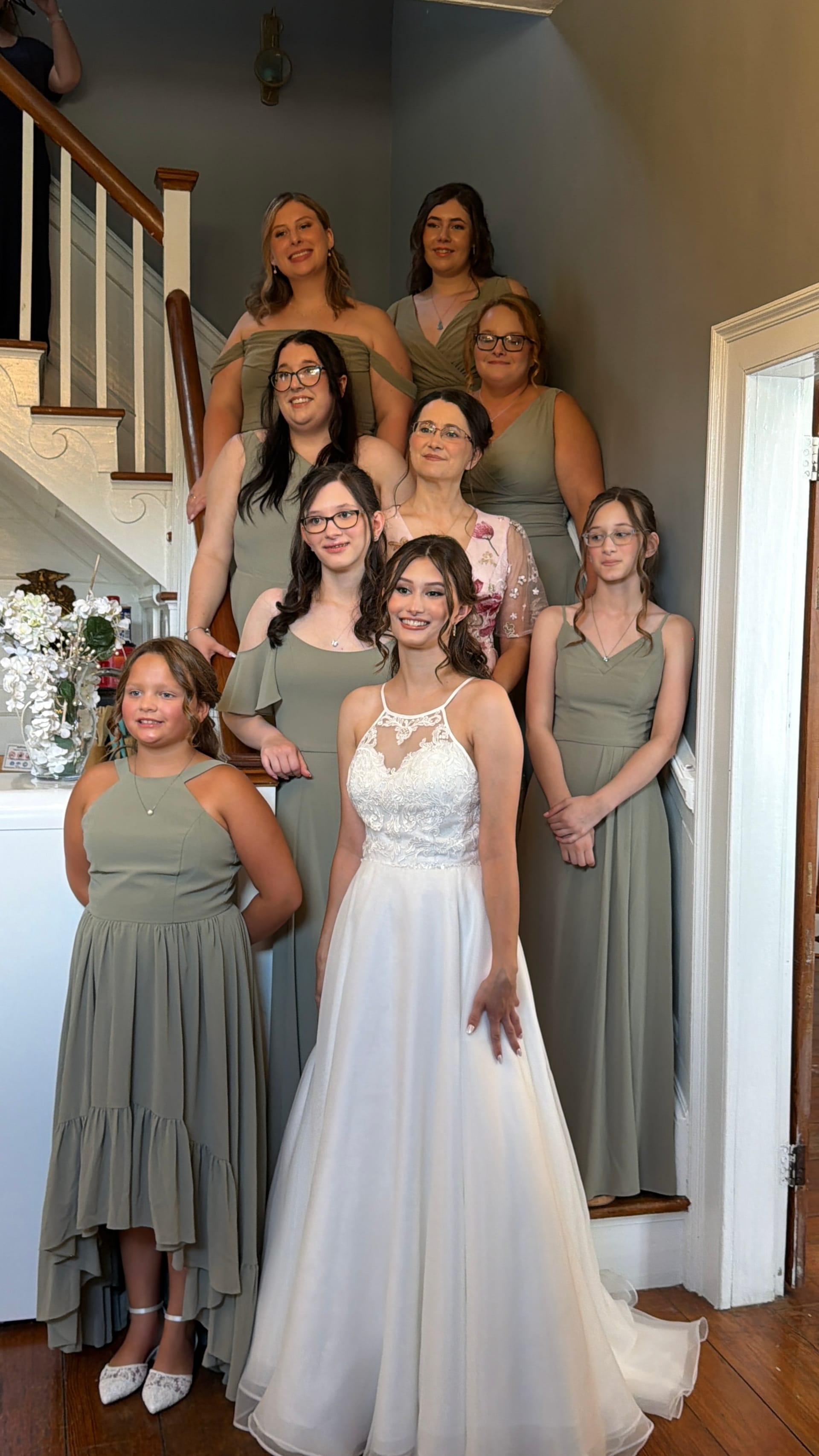 Bride in a white lace halter gown stands in front of bridesmaids and junior attendants in sage green dresses arranged on an interior staircase before the wedding ceremony.