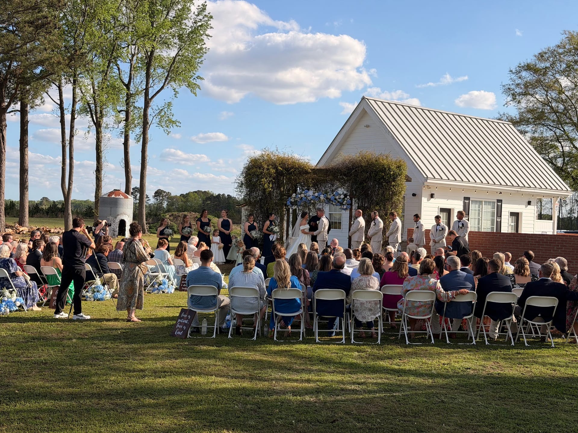 Guests seated on a sunny ceremony lawn watch a wedding party gathered beneath a floral arch in front of a white chapel at Scarborough House estate.