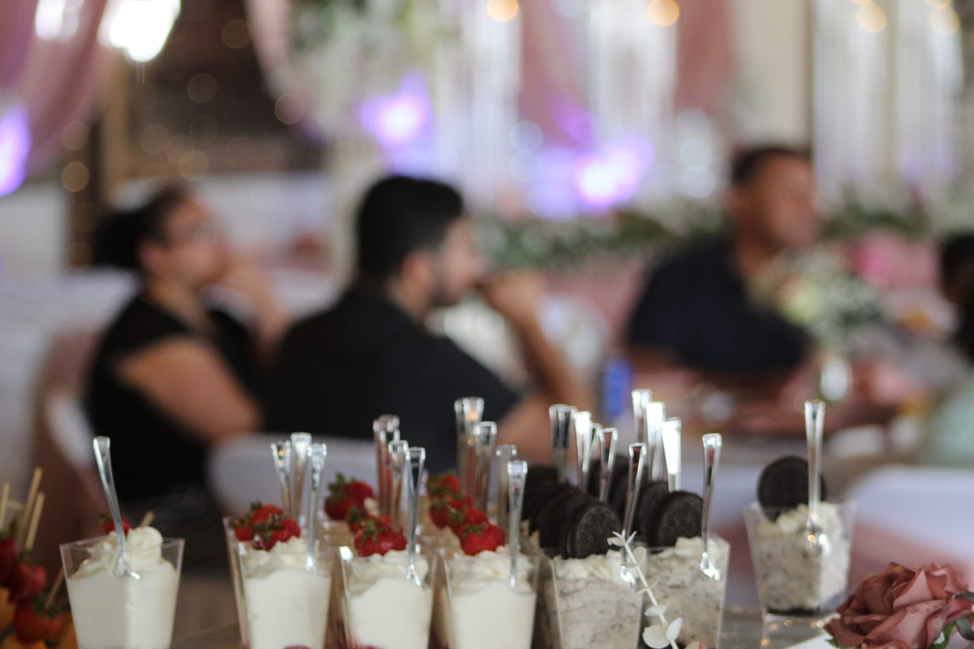 Elegant wedding dessert display featuring individual cream cups topped with fresh strawberries and Oreo cookies with silver spoons, set against a softly blurred grand hall reception backdrop with pink drapery and purple lighting.