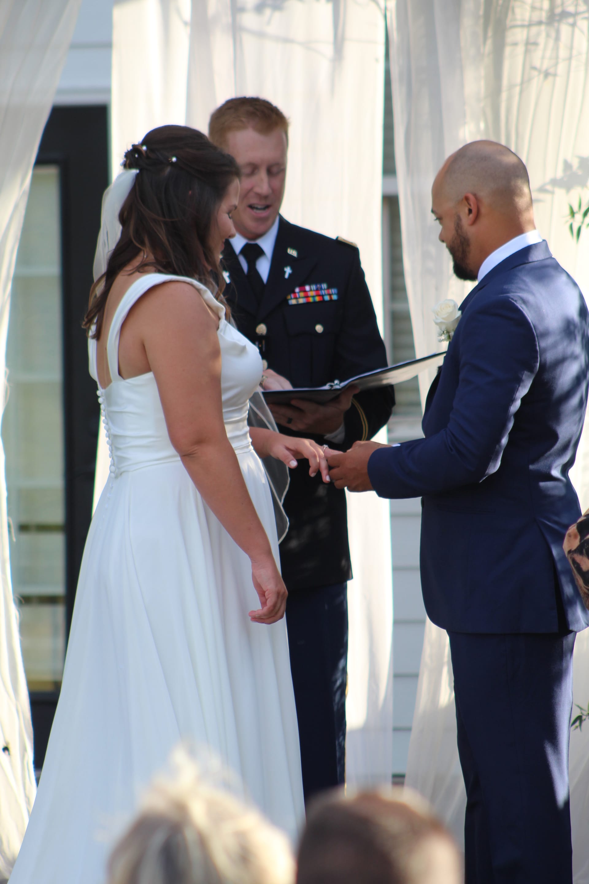 Bride and groom exchange rings during an outdoor wedding ceremony officiated by a uniformed military officer beneath a draped white archway at Scarborough House.