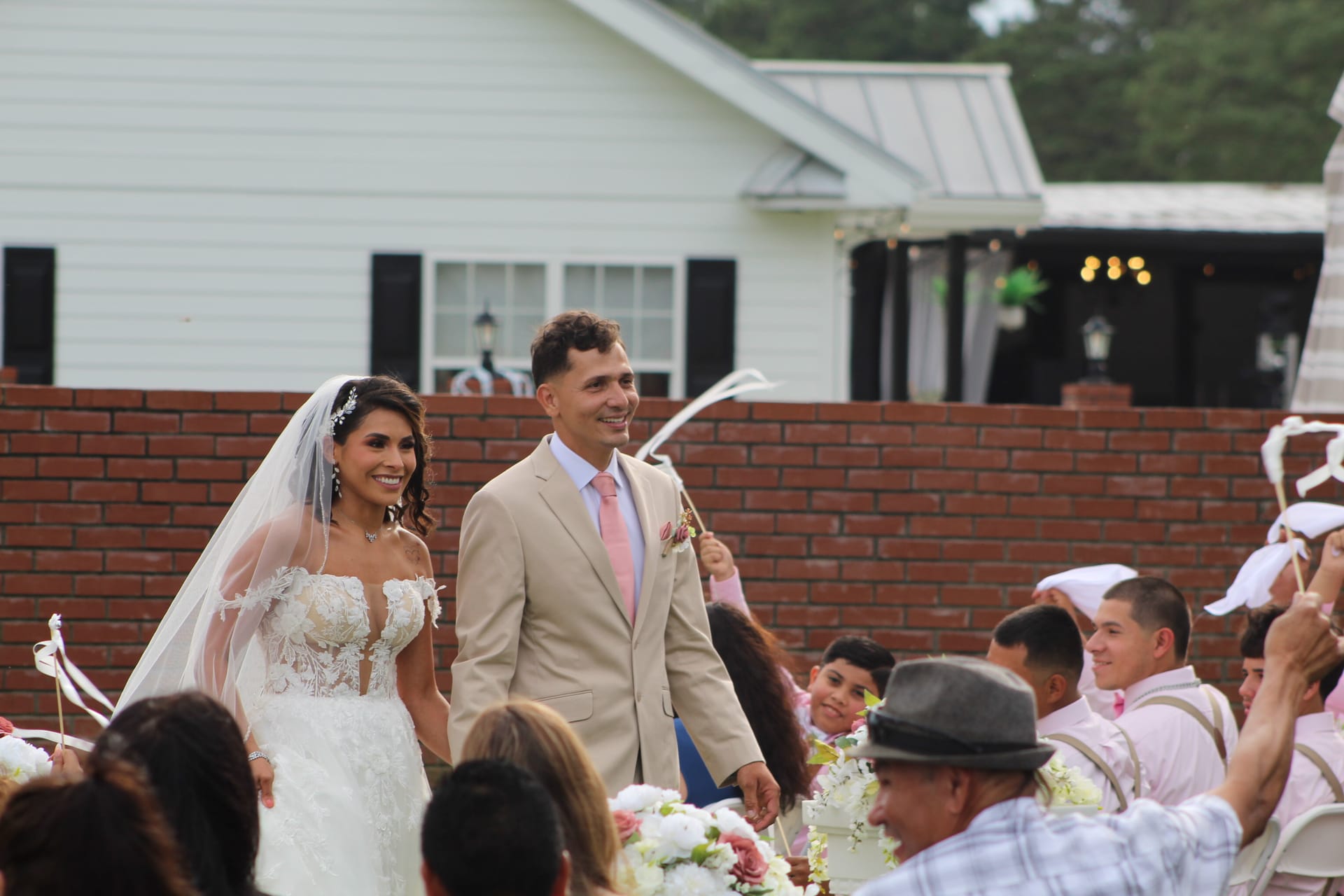 Smiling bride in lace gown and groom in tan suit walk the recessional aisle as guests wave ribbon wands at an outdoor wedding at Scarborough House estate.