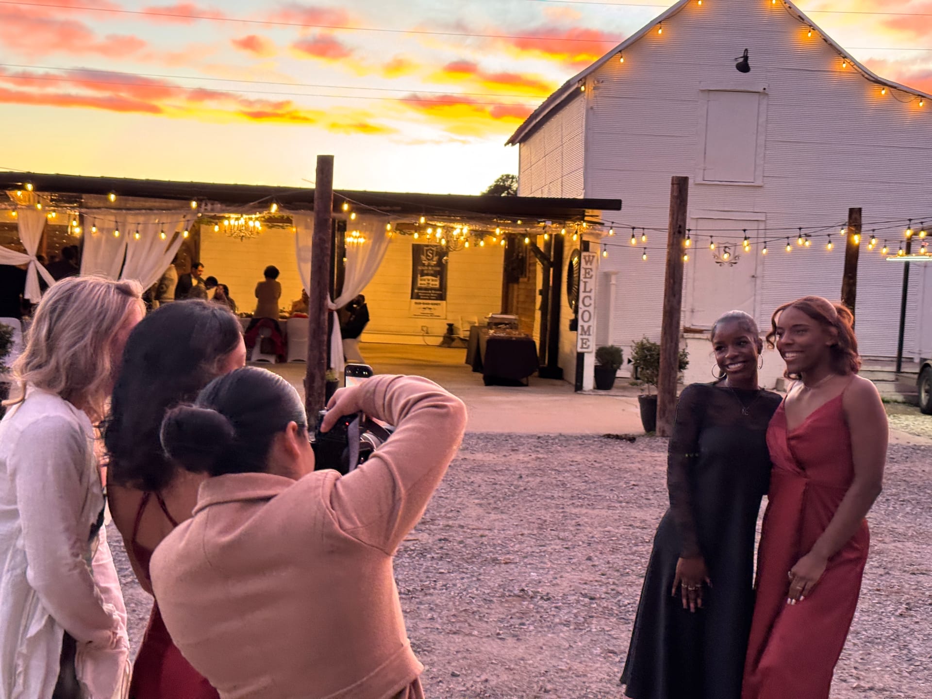 Two guests in formal black and red gowns pose outside the white barn at Scarborough House, with string lights and a sunset sky.