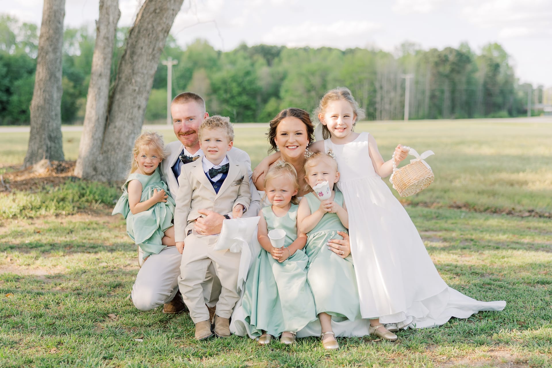 Bride and groom kneeling on grass beneath oak trees with four young children in mint green dresses and a tan suit during a sunny outdoor wedding at Scarborough House.