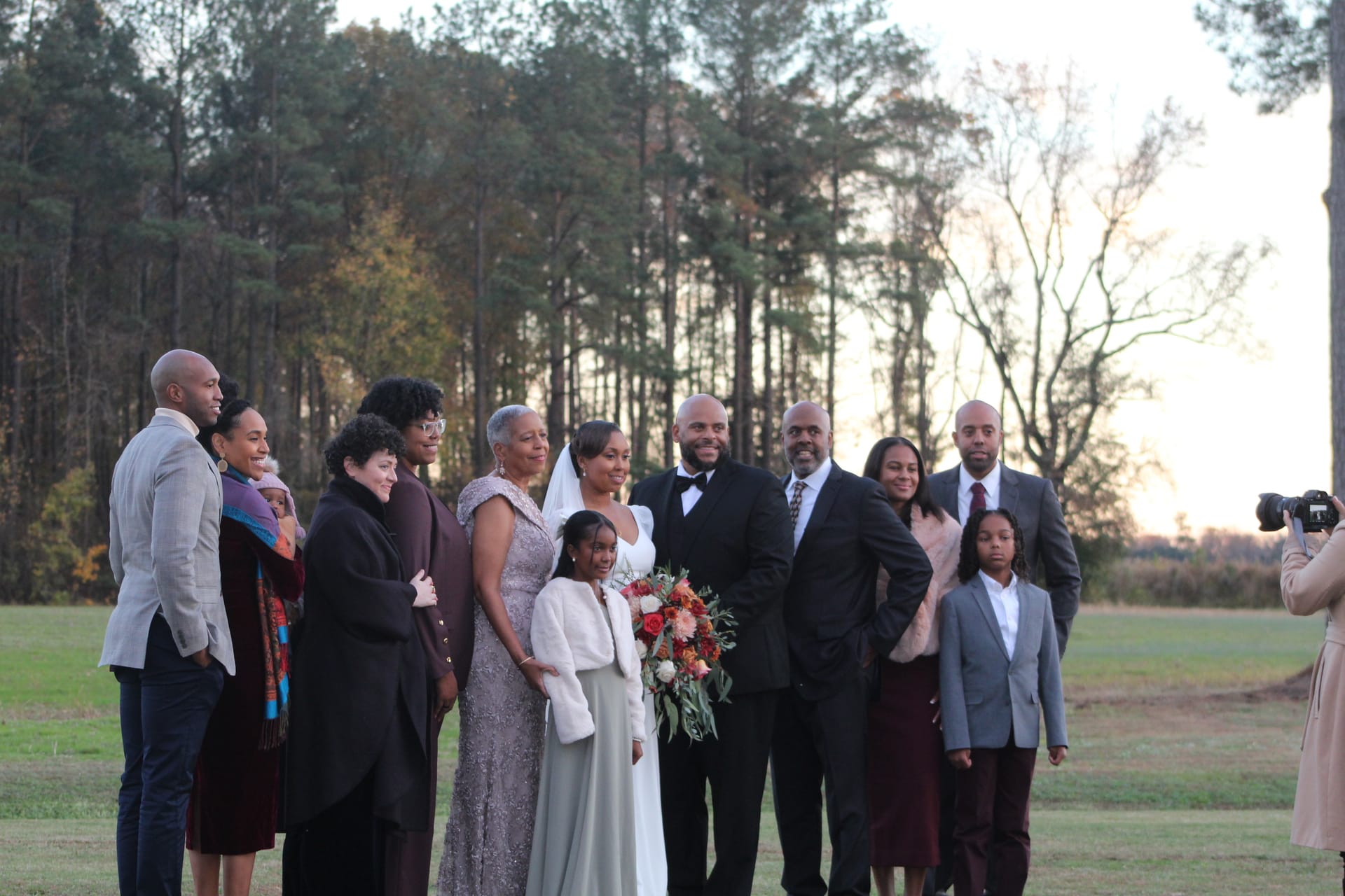 Bride and groom pose with family members for a group portrait on an open lawn at sunset surrounded by tall pine trees at a historic estate wedding venue.