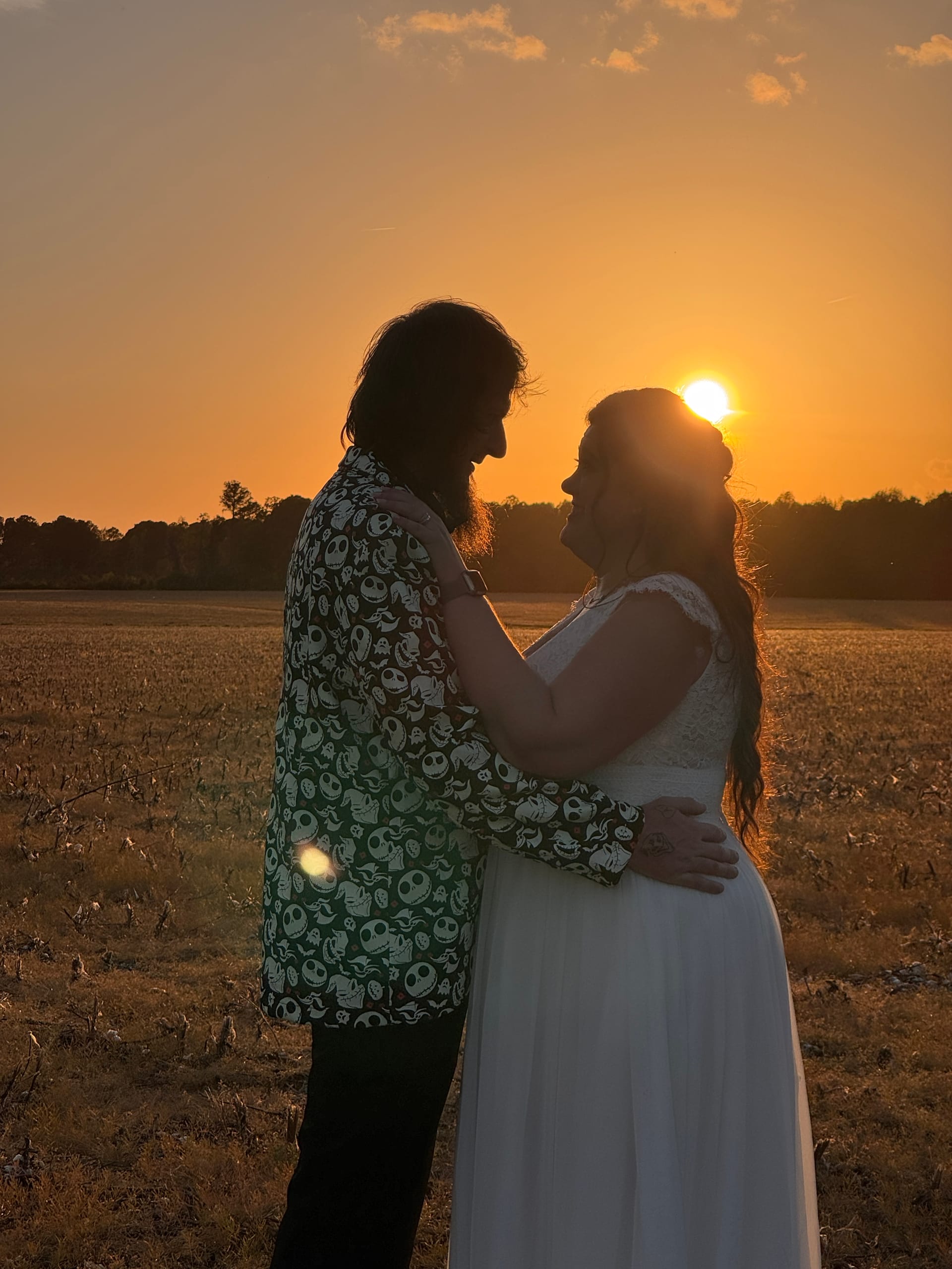 Bride in white gown and groom in a patterned shirt share a romantic embrace silhouetted against a glowing orange sunset over the open farmland fields at Scarborough House.