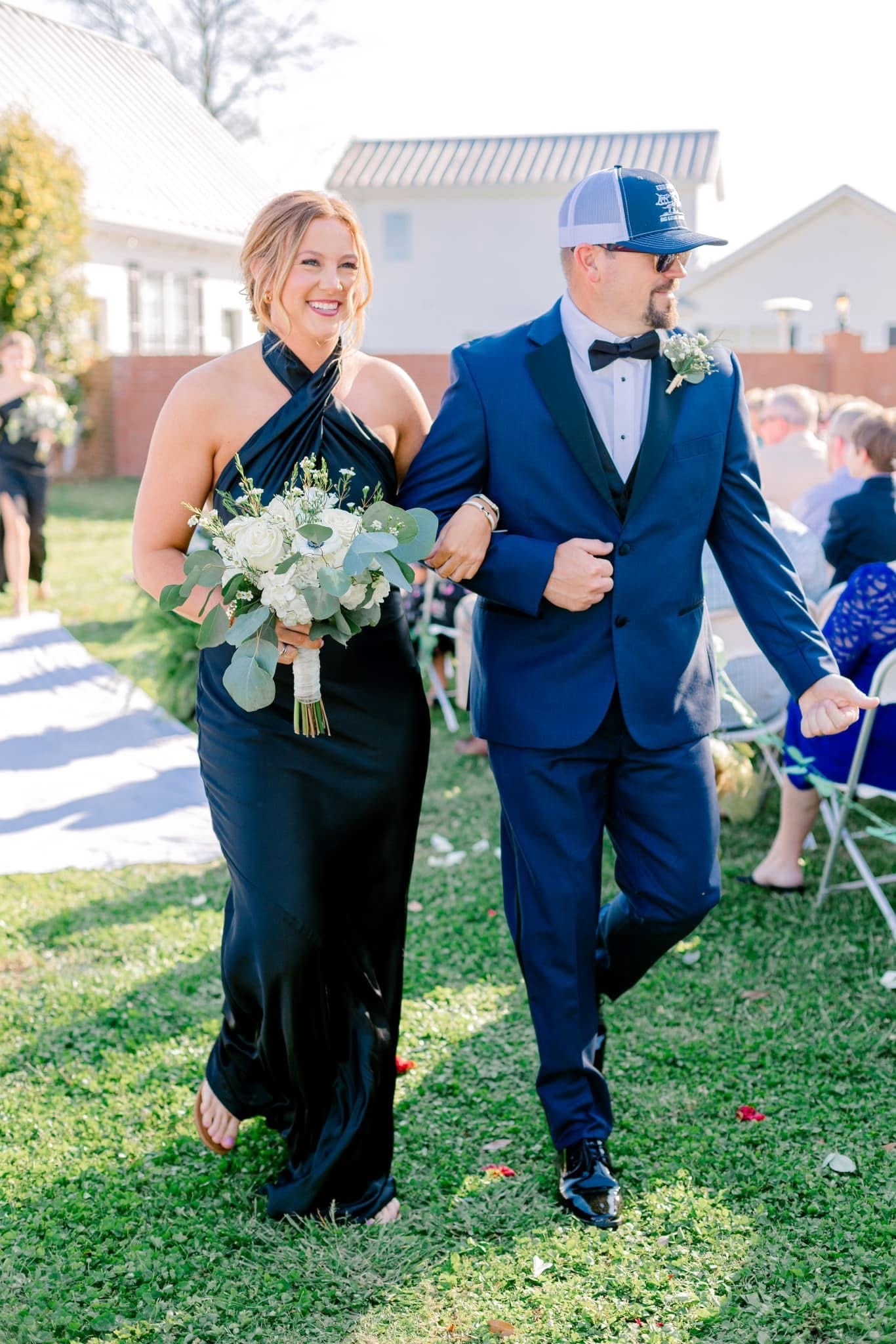 A smiling bridesmaid in a black halter gown carrying a white and greenery bouquet walks arm-in-arm with a groomsman in a navy tuxedo down the outdoor ceremony aisle at Scarborough House.