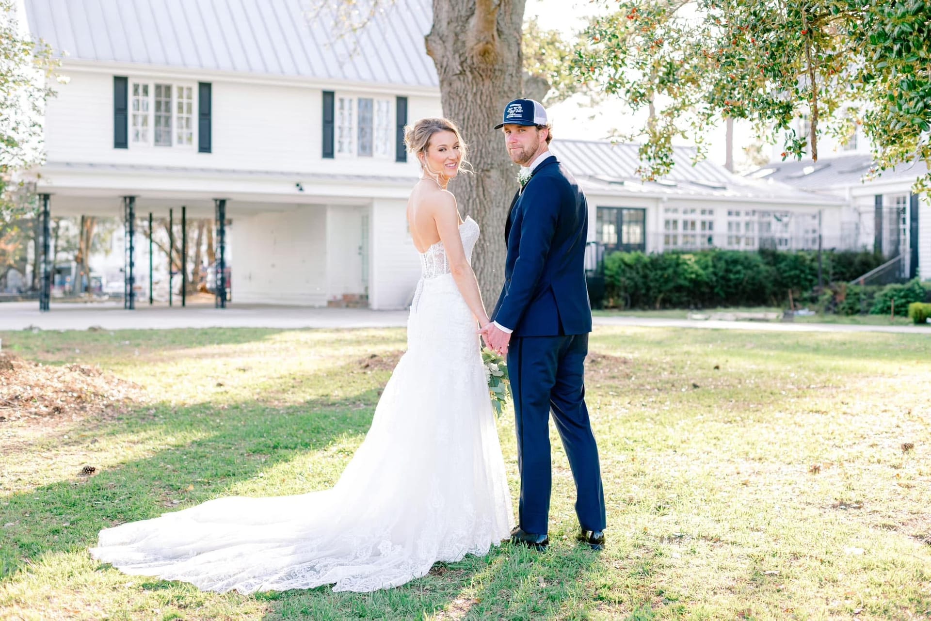 Bride in a lace sweetheart gown and groom in a navy suit hold hands and look back over their shoulders while standing on the sunlit lawn in front of the historic white Scarborough House manor.