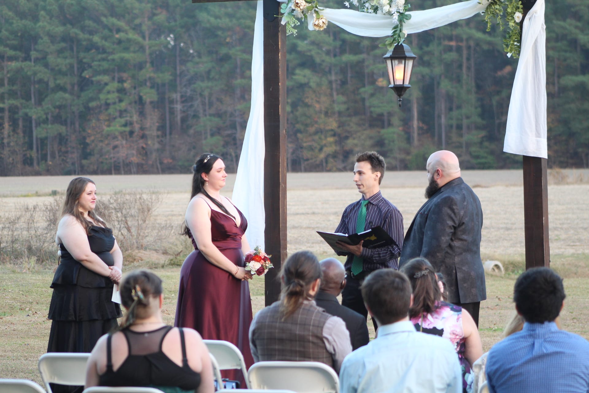 An outdoor wedding ceremony at dusk beneath a wooden arch draped with white fabric and floral greenery, with a bridesmaid in burgundy, officiant, and groom gathered as guests look on across an open field.