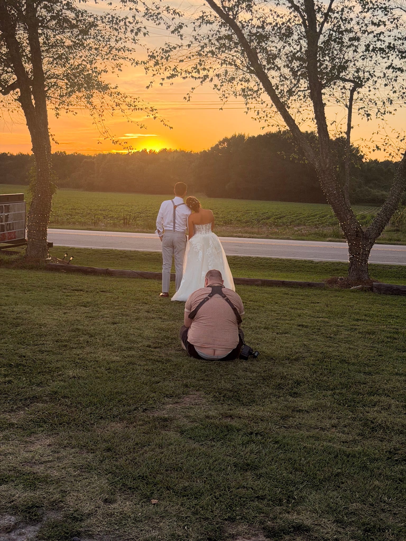 A bride in a white gown and groom in suspenders stand together facing a vivid orange sunset over open farmland fields while their wedding photographer crouches on the grass to capture the moment.