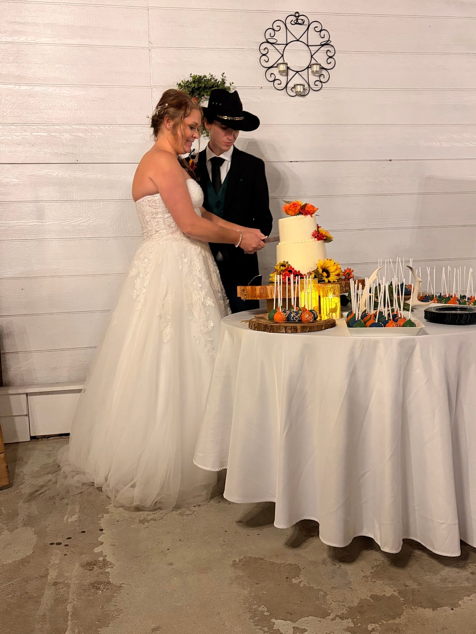 Bride in a strapless lace ball gown and groom in a black suit and cowboy hat cut their two-tier white wedding cake adorned with fall flowers at a rustic autumn wedding reception.