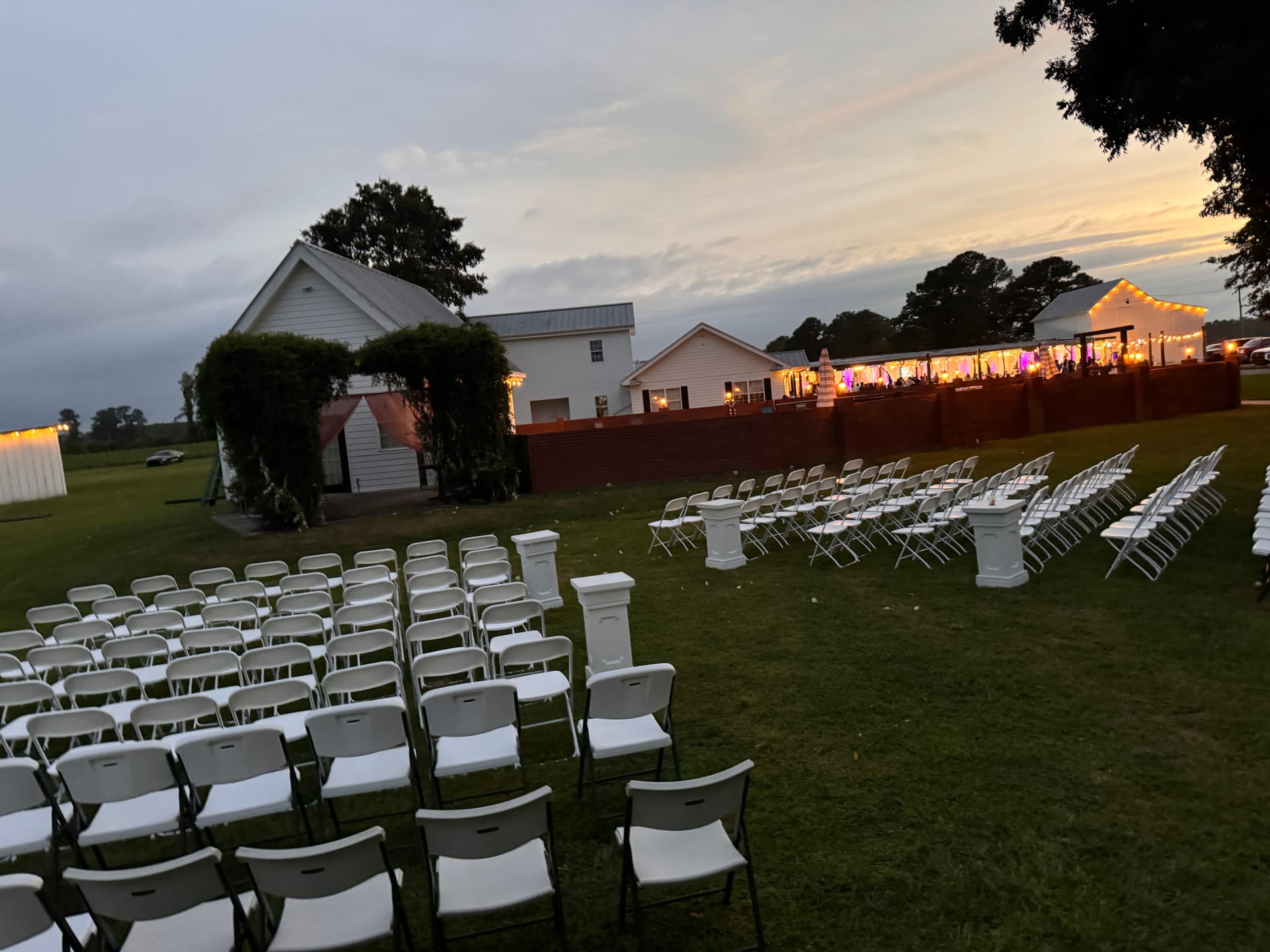 Rows of white folding chairs arranged on the ceremony lawn at dusk at Scarborough House with warm reception lights glowing from the pavilion in the background at sunset.