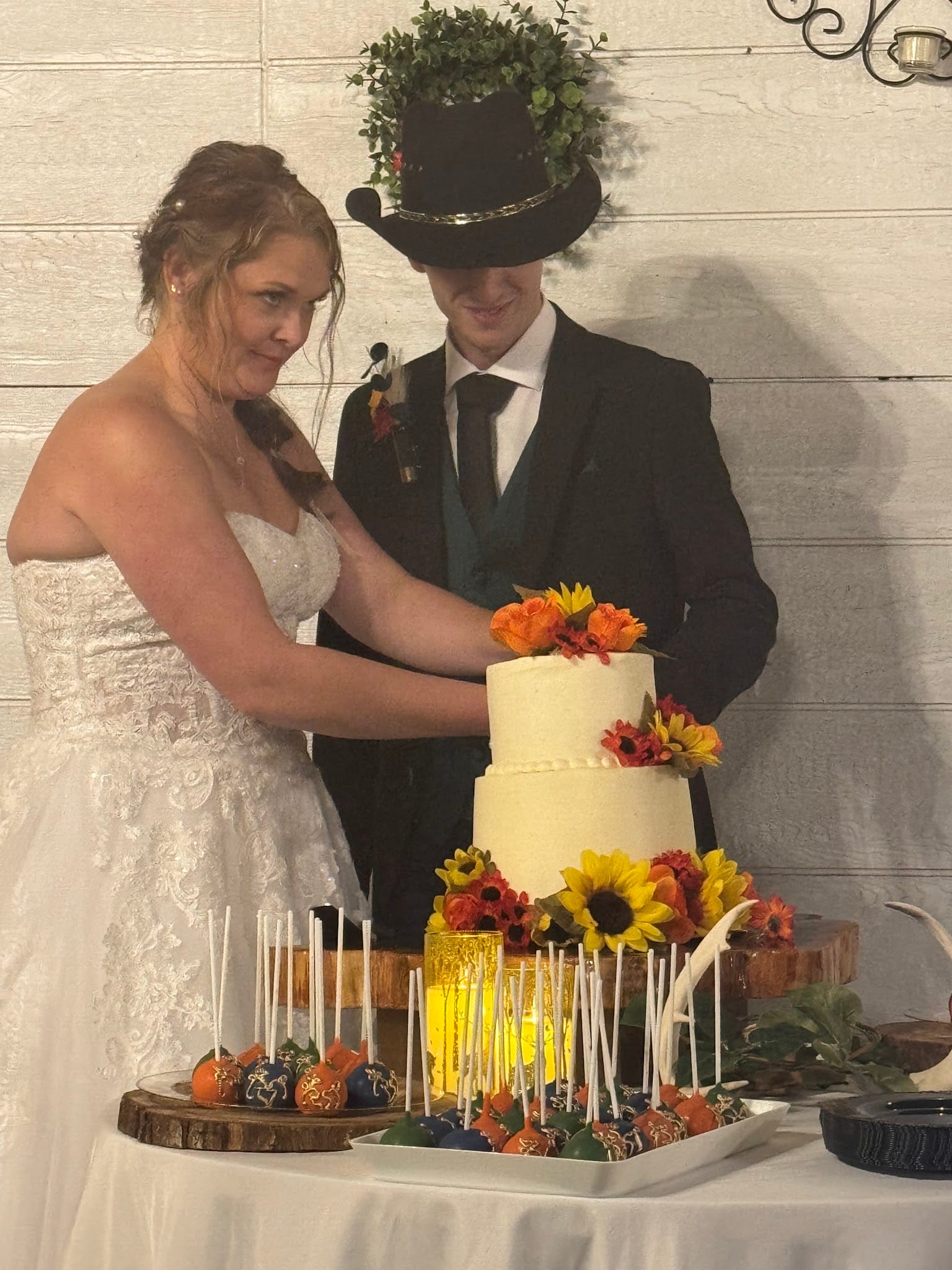 Bride in lace gown and groom in black suit with decorated top hat cut their two-tier white wedding cake adorned with sunflowers and autumn blooms at Scarborough House.