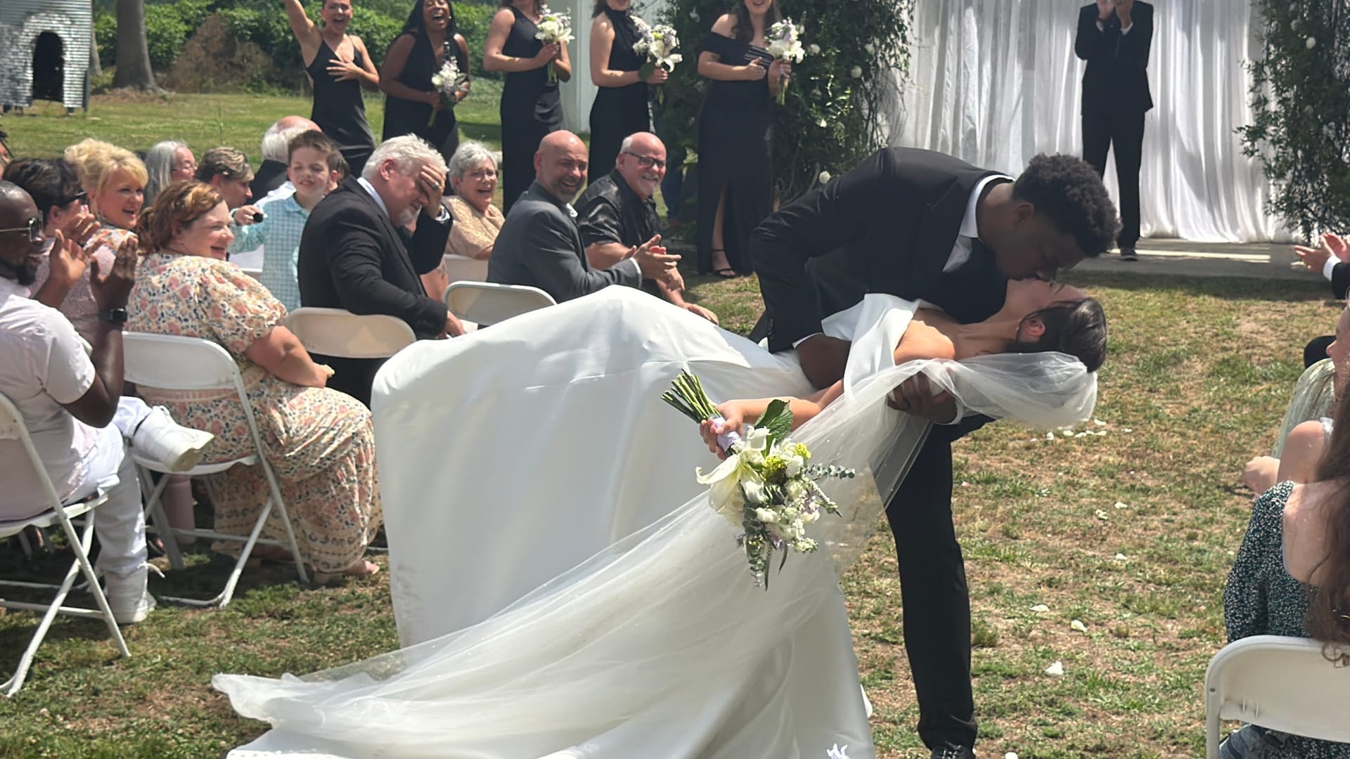 Groom dramatically dips and kisses his bride mid-aisle during their outdoor wedding ceremony at Scarborough House as joyful guests cheer and applaud around them.