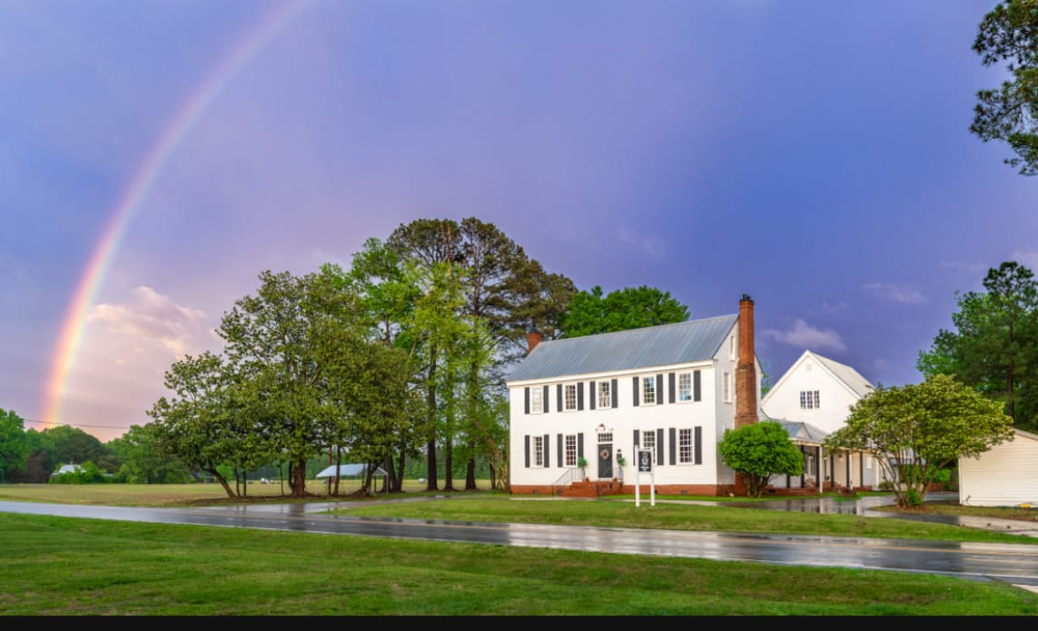A rainbow arches over the white Scarborough House manor and surrounding trees, with a wet road in the foreground.