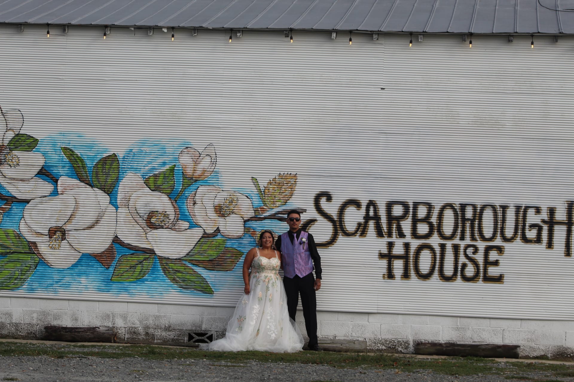Bride in a floral ballgown and groom in a purple vest pose together in front of the Scarborough House magnolia mural on the exterior barn wall.