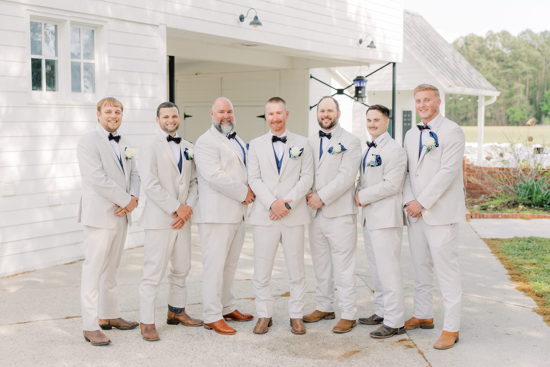 Groom and six groomsmen in matching light grey suits with navy bow ties and boutonnieres pose together outside the white barn facade at Scarborough House.