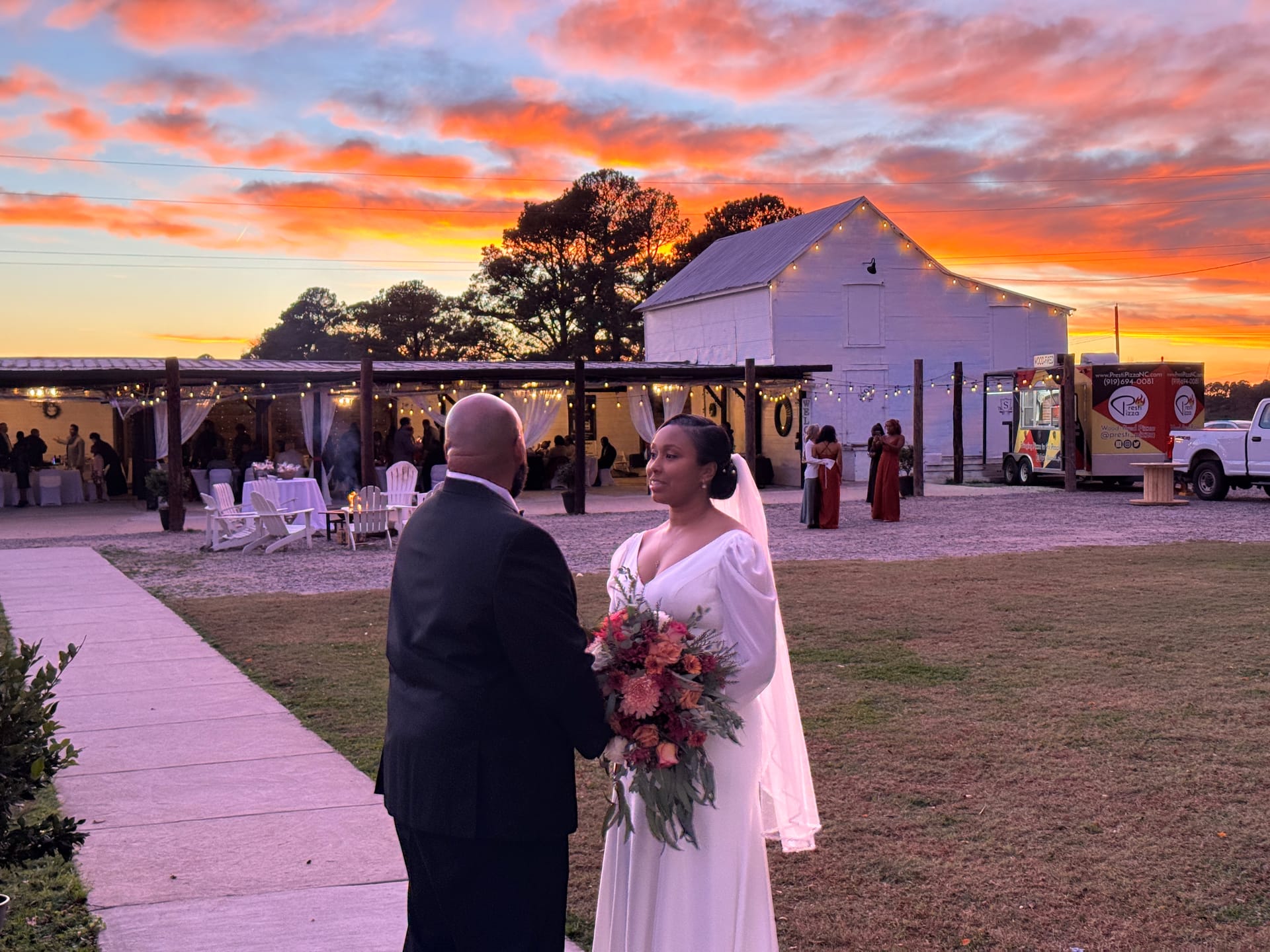 Bride in a flowing white gown holding a colorful bouquet faces her groom in a black suit at dusk with a dramatic orange and pink sunset sky over a white barn wedding venue.