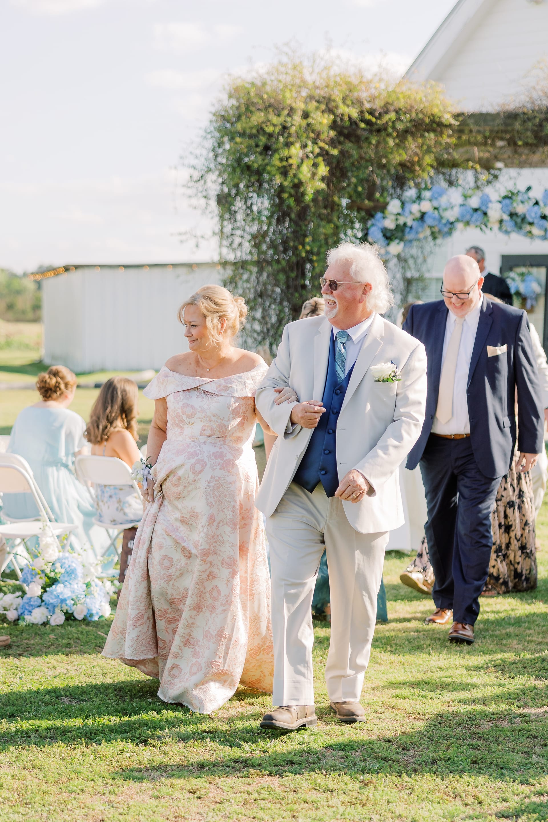 A smiling couple dressed in formal attire walk arm-in-arm down the ceremony lawn aisle at Scarborough House, with blue hydrangea floral arrangements and a vine-covered white barn in the background.