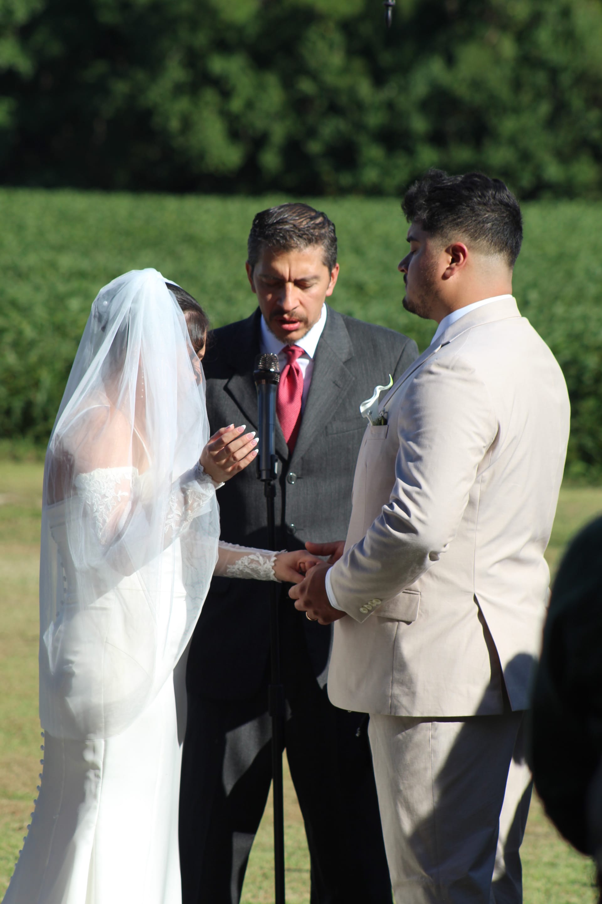 Bride in white lace gown and veil holds hands with groom in tan suit as officiant speaks during an outdoor wedding ceremony on the estate lawn surrounded by lush greenery.