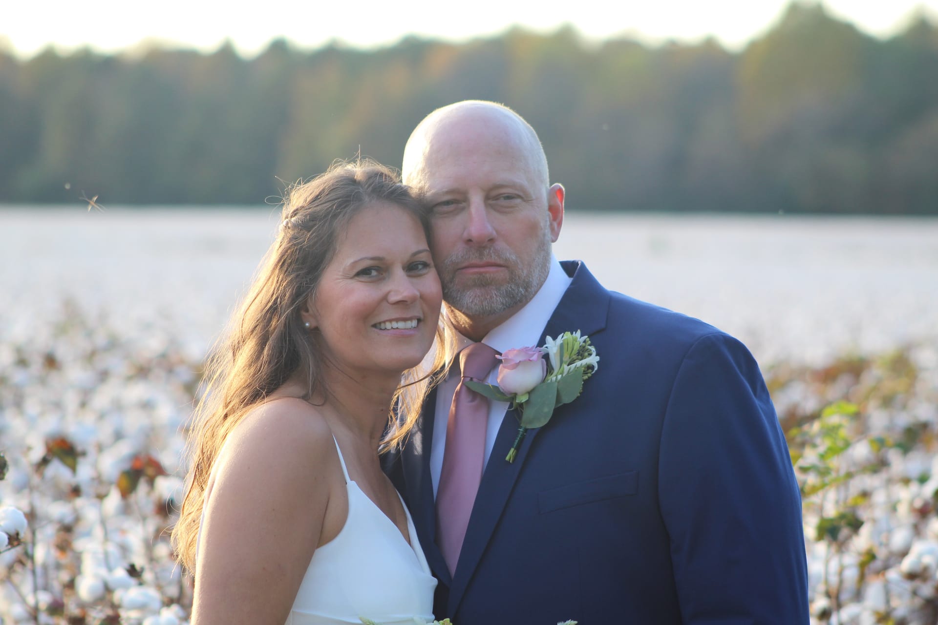 Bride in white dress and groom in navy suit with pink rose boutonniere pose together in a cotton field at golden hour with a treeline in the background.