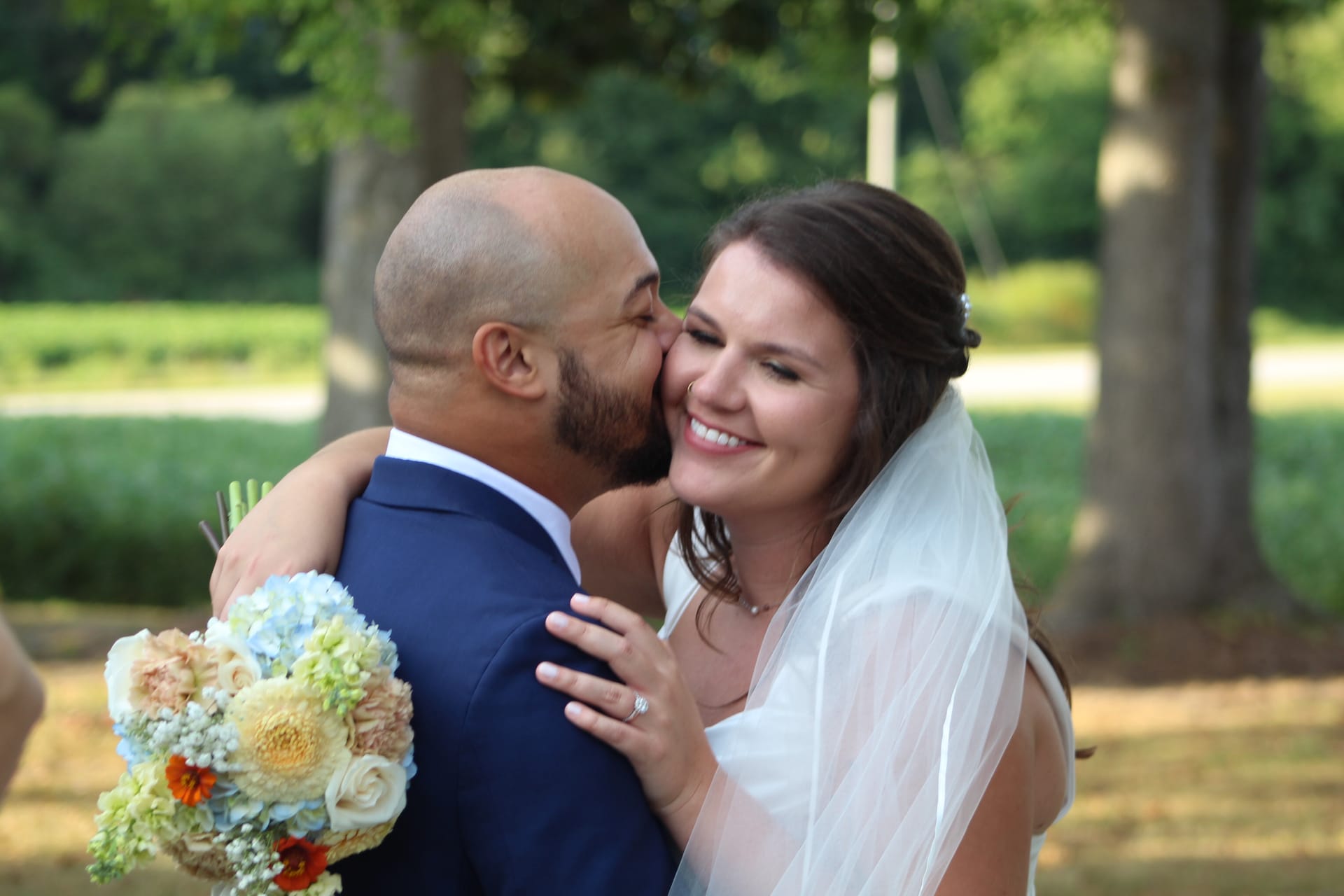 Bride smiles radiantly while holding a pastel bouquet as groom kisses her cheek beneath the oak trees at Scarborough House estate.