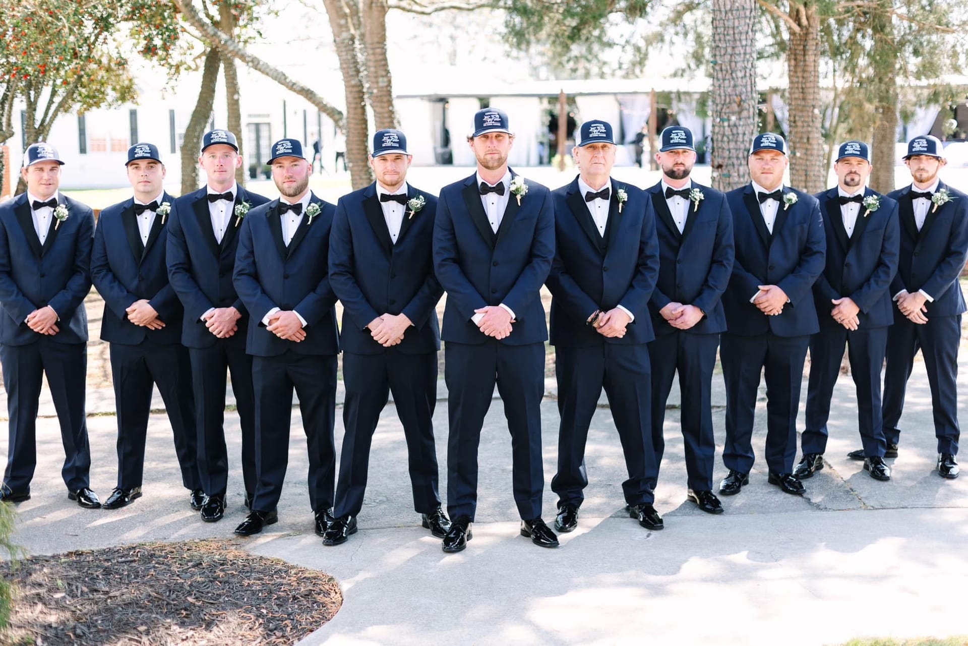 Groom and ten groomsmen dressed in matching navy tuxedos with bow ties, boutonnieres, and coordinating baseball caps stand in a row on the ceremony grounds at Scarborough House surrounded by oak trees.